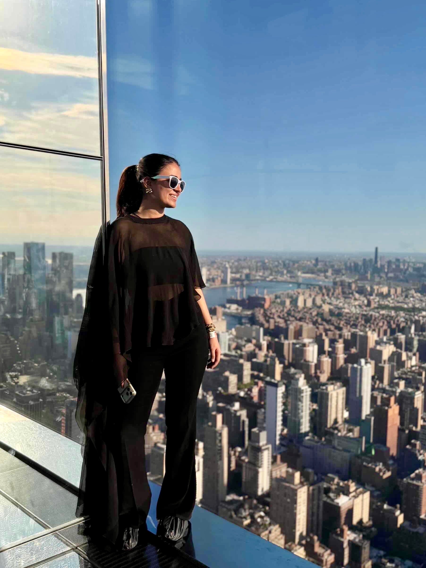 A woman wearing a Khamna'S vRINDA CAPE SET s stands on a high-rise balcony overlooking a dense city skyline with a river and blue sky.