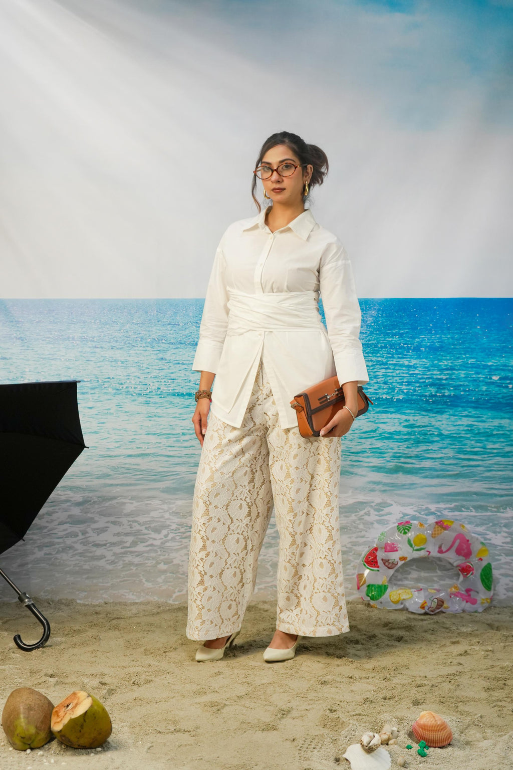 A woman stands in a beach-themed studio setting wearing a white long-sleeve collared shirt and matching cream lace wide-leg pants, holding a brown clutch. The background shows blue ocean water and sky, with props including coconuts, a ring float, and a black umbrella.