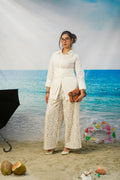 A woman stands in a beach-themed studio setting wearing a white long-sleeve collared shirt and matching cream lace wide-leg pants, holding a brown clutch. The background shows blue ocean water and sky, with props including coconuts, a ring float, and a black umbrella.
