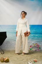 A woman stands in a beach-themed studio setting wearing a white long-sleeve collared shirt and matching cream lace wide-leg pants, holding a brown clutch. The background shows blue ocean water and sky, with props including coconuts, a ring float, and a black umbrella.