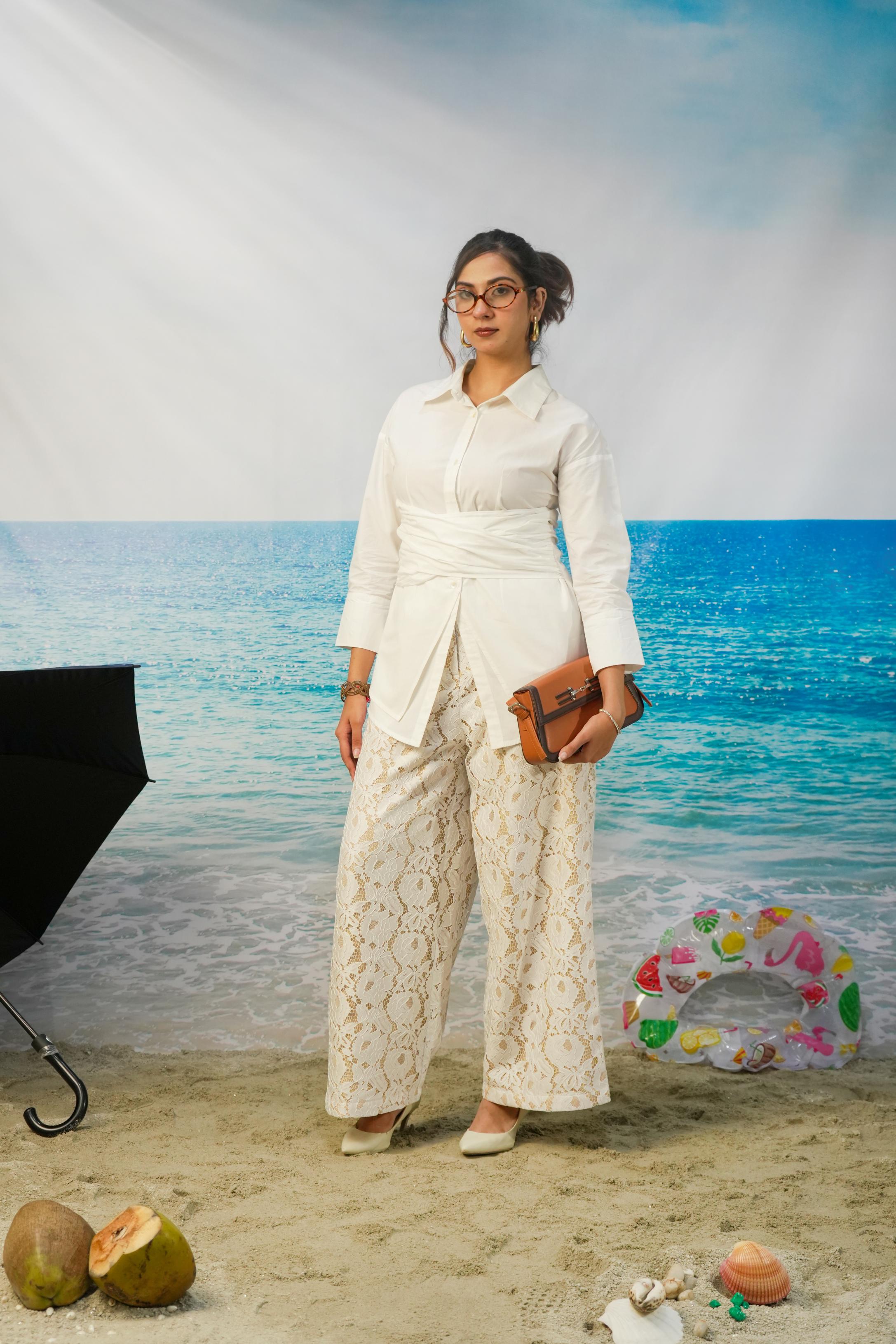 A woman stands in a beach-themed studio setting wearing a white long-sleeve collared shirt and matching cream lace wide-leg pants, holding a brown clutch. The background shows blue ocean water and sky, with props including coconuts, a ring float, and a black umbrella.
