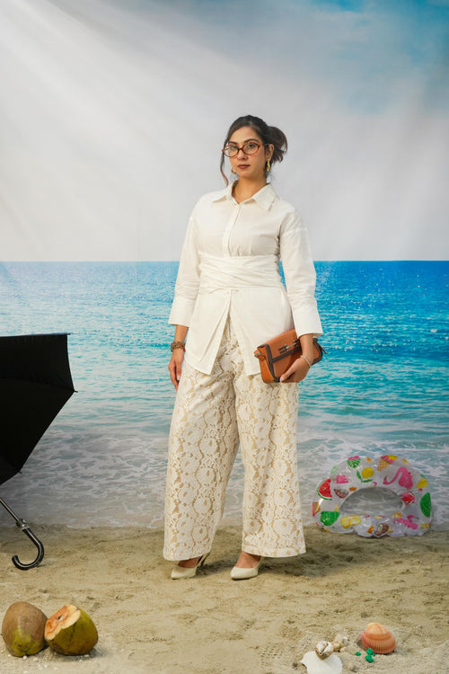 A woman stands in a beach-themed studio setting wearing a white long-sleeve collared shirt and matching cream lace wide-leg pants, holding a brown clutch. The background shows blue ocean water and sky, with props including coconuts, a ring float, and a black umbrella.