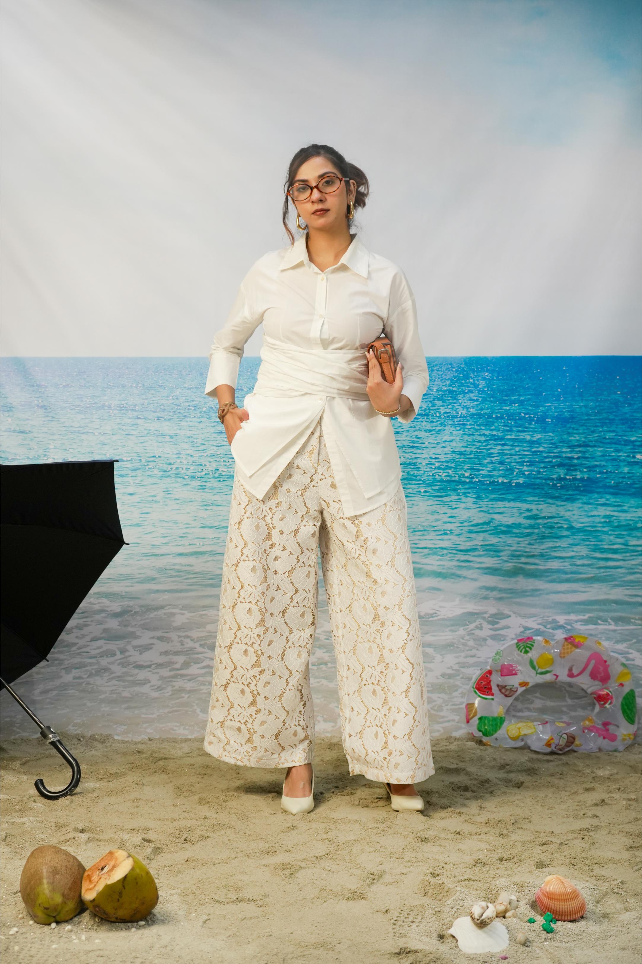 A woman modeling a white long-sleeve button-up shirt and matching white lace wide-leg pants, standing on a sandy beach backdrop with ocean scenery behind her. Props include a black umbrella on the left, a colorful float on the right, and coconuts and shells on the sand.