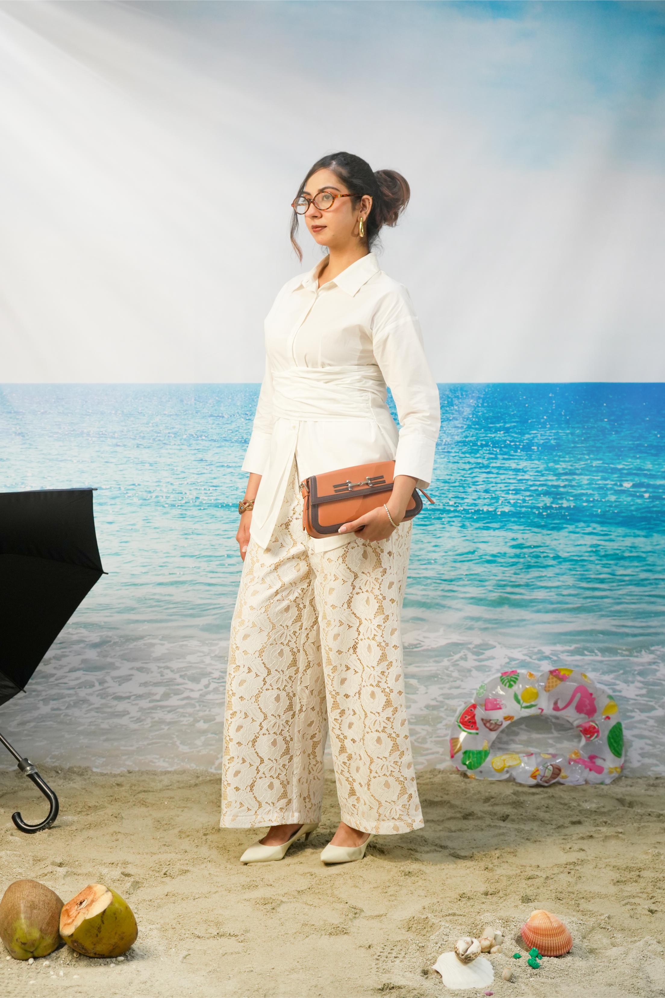 A woman wearing a cream lace long-sleeve top and matching wide-leg pants stands on a beach backdrop holding a brown leather clutch. The scene includes a beach umbrella, coconuts, a swim ring, and a printed ocean-and-sky background.