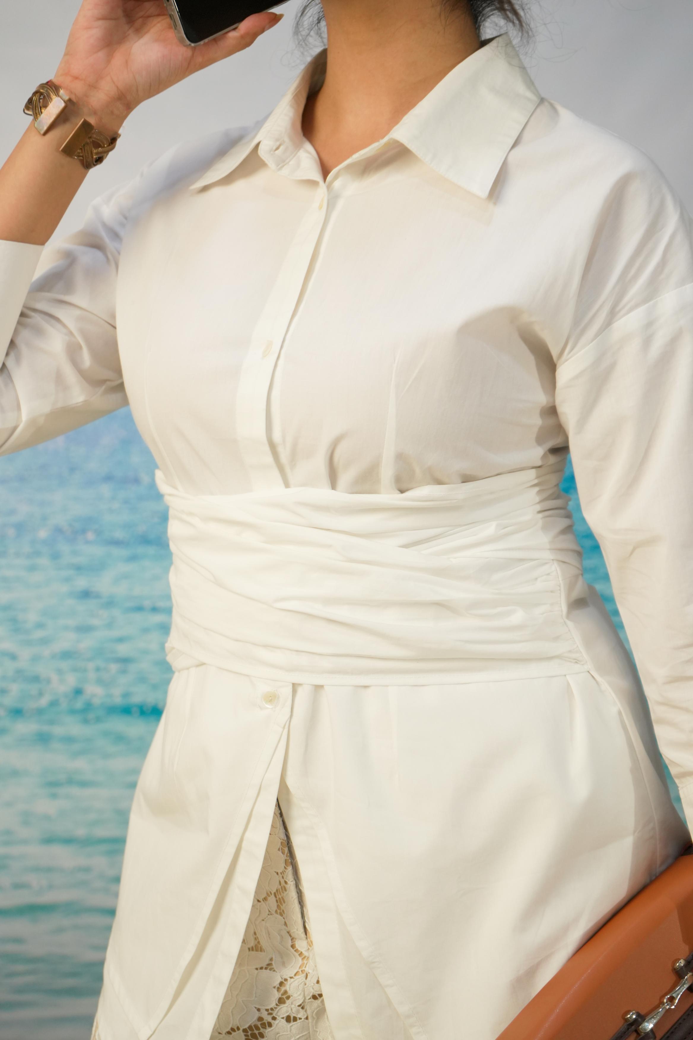 A woman wearing a cream-colored long-sleeve collared shirt dress with a ruched waist, standing in front of a blue ocean background.