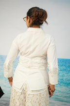 A woman viewed from behind wearing a white long-sleeve collared shirt with a gathered waist and a cream lace skirt, standing by the ocean.