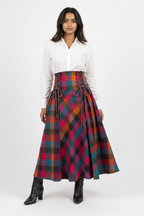 A model wearing a white long-sleeve button-up shirt and a high-waisted, pleated A-line skirt in a vibrant red, orange, blue, and green tartan plaid pattern. The skirt features a lace-up corset waist.