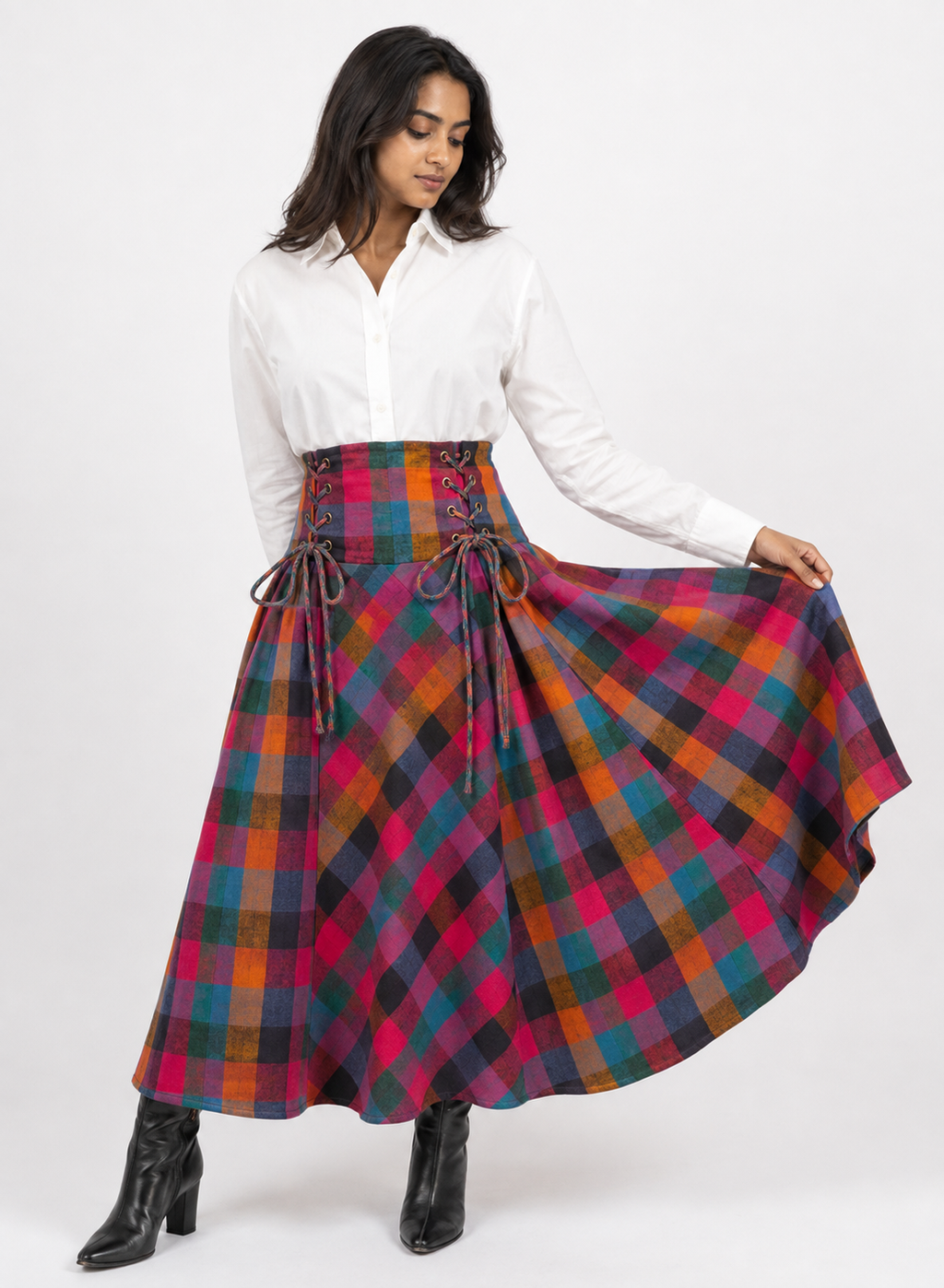 A woman modeling a high-waisted, full-length flared skirt in a vibrant multicolor tartan plaid pattern. The skirt features a lace-up corset waistband.
