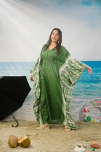 A woman modeling a green kaftan dress with wide patterned sleeves, standing on a sandy beach set with a blue ocean backdrop, props including a black umbrella, coconuts, and a swim ring.