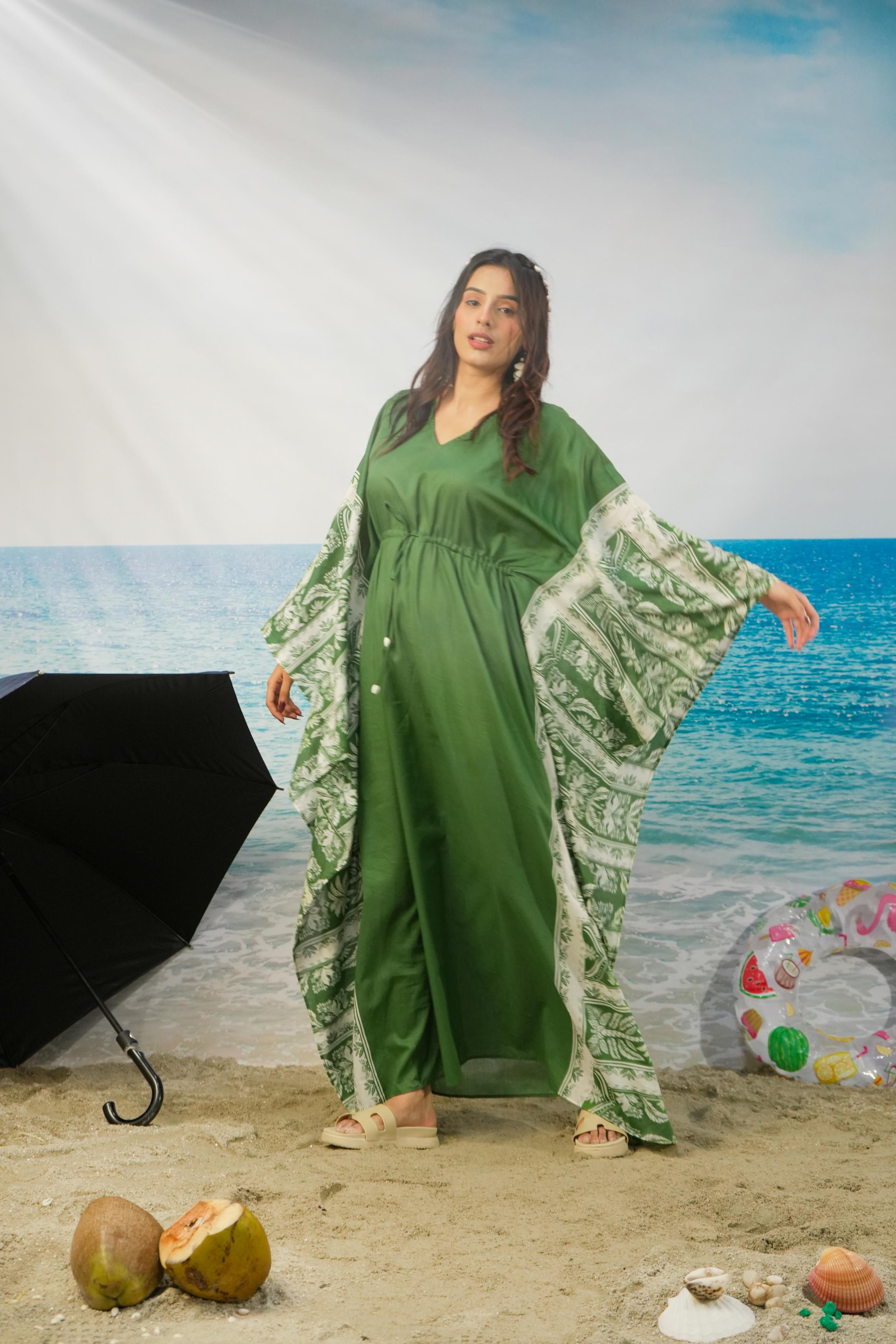 A woman modeling a green kaftan dress with wide patterned sleeves, standing on a sandy beach set with a blue ocean backdrop, props including a black umbrella, coconuts, and a swim ring.