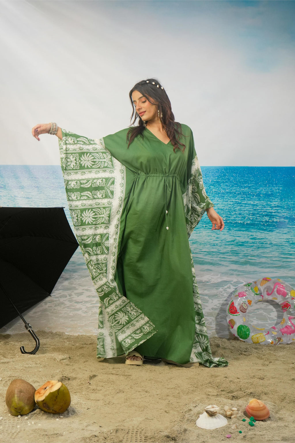 A woman modeling a green kaftan dress with wide sleeves and a matching printed dupatta, posed on a beach backdrop with sand, ocean, coconuts, and a black umbrella visible.