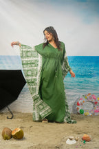 A woman modeling a green kaftan dress with wide sleeves and a matching printed dupatta, posed on a beach backdrop with sand, ocean, coconuts, and a black umbrella visible.