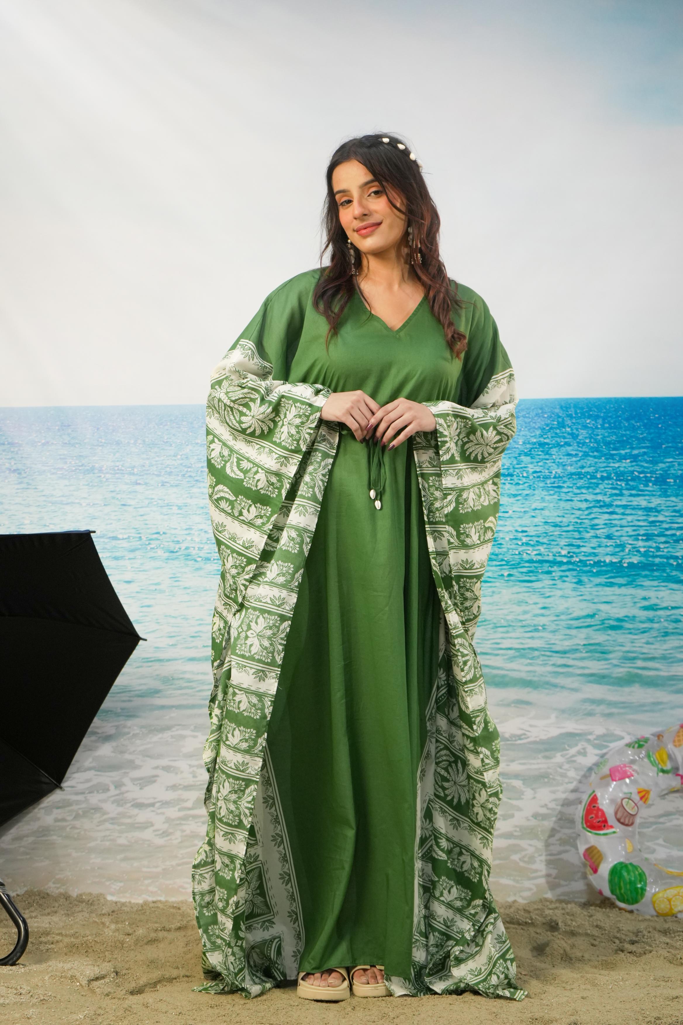 A woman stands on a beach backdrop wearing a flowing sage green dress with wide sleeves that have a white and green patterned border. Studio lighting equipment is visible on the left, and a beach ball is on the right.