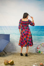 A woman stands on a beach backdrop wearing a colorful plaid midi dress with three-quarter sleeves and a square neckline, viewed from behind. The dress has a bold plaid pattern in red, blue, yellow, and black.