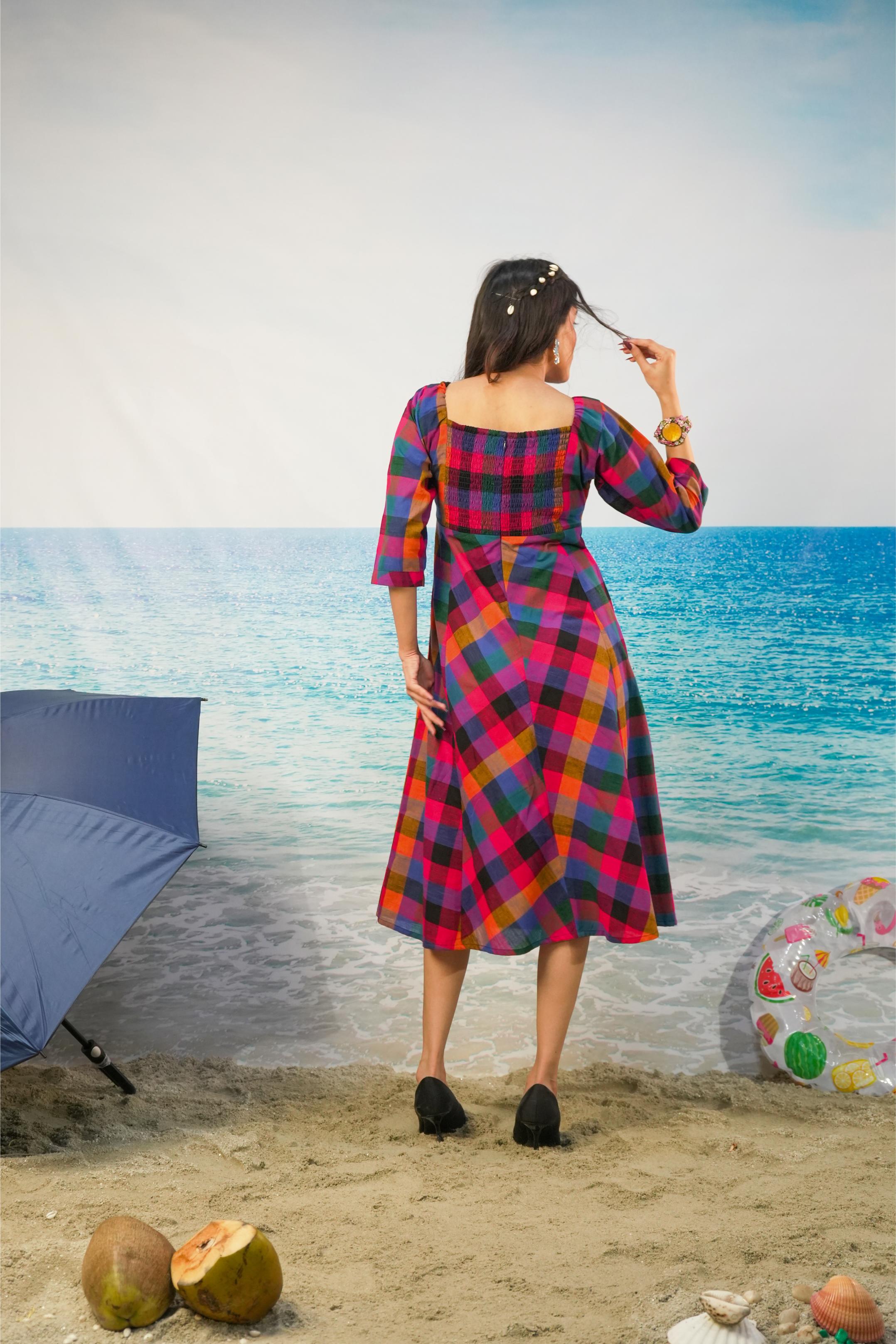 A woman stands on a beach backdrop wearing a colorful plaid midi dress with three-quarter sleeves and a square neckline, viewed from behind. The dress has a bold plaid pattern in red, blue, yellow, and black.