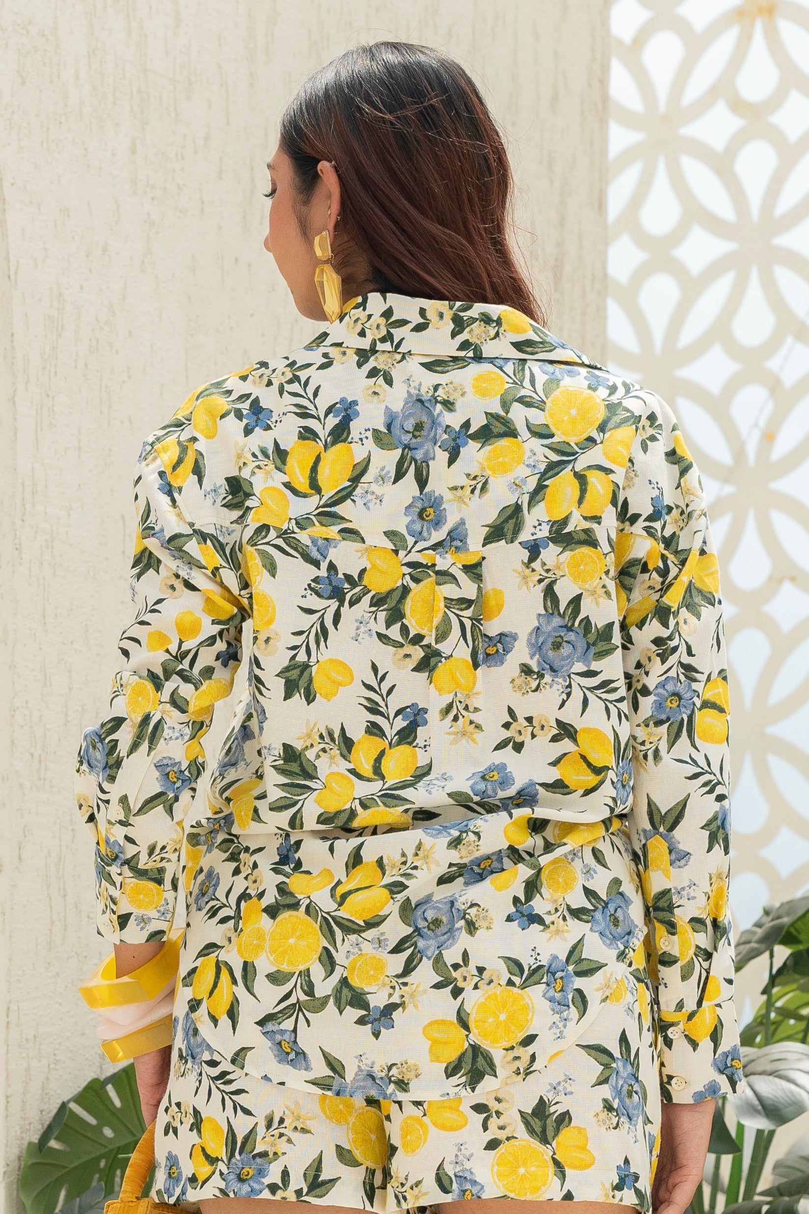 Woman in a Citrus Bloom Oversized Shirt – Printed Cotton standing on a staircase with decorative white walls and plants.
