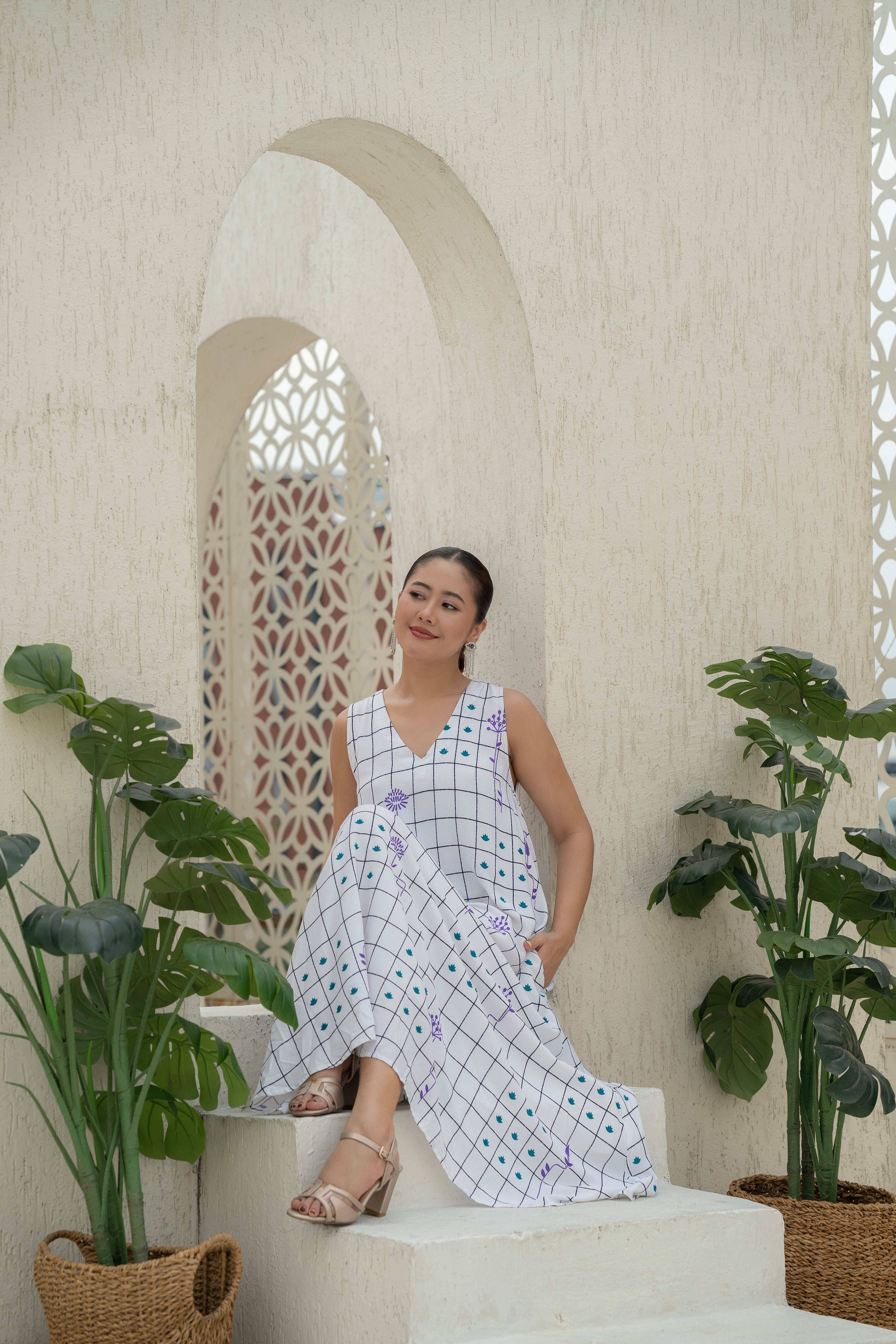 Woman in a checkered dress sitting on a white bench with plants around her.