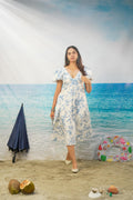 A woman stands on a beach-themed backdrop wearing a white midi dress with a blue floral print and puff sleeves. The scene includes sand, ocean water, a closed navy umbrella, a colorful inflatable ring, coconuts, and shells.