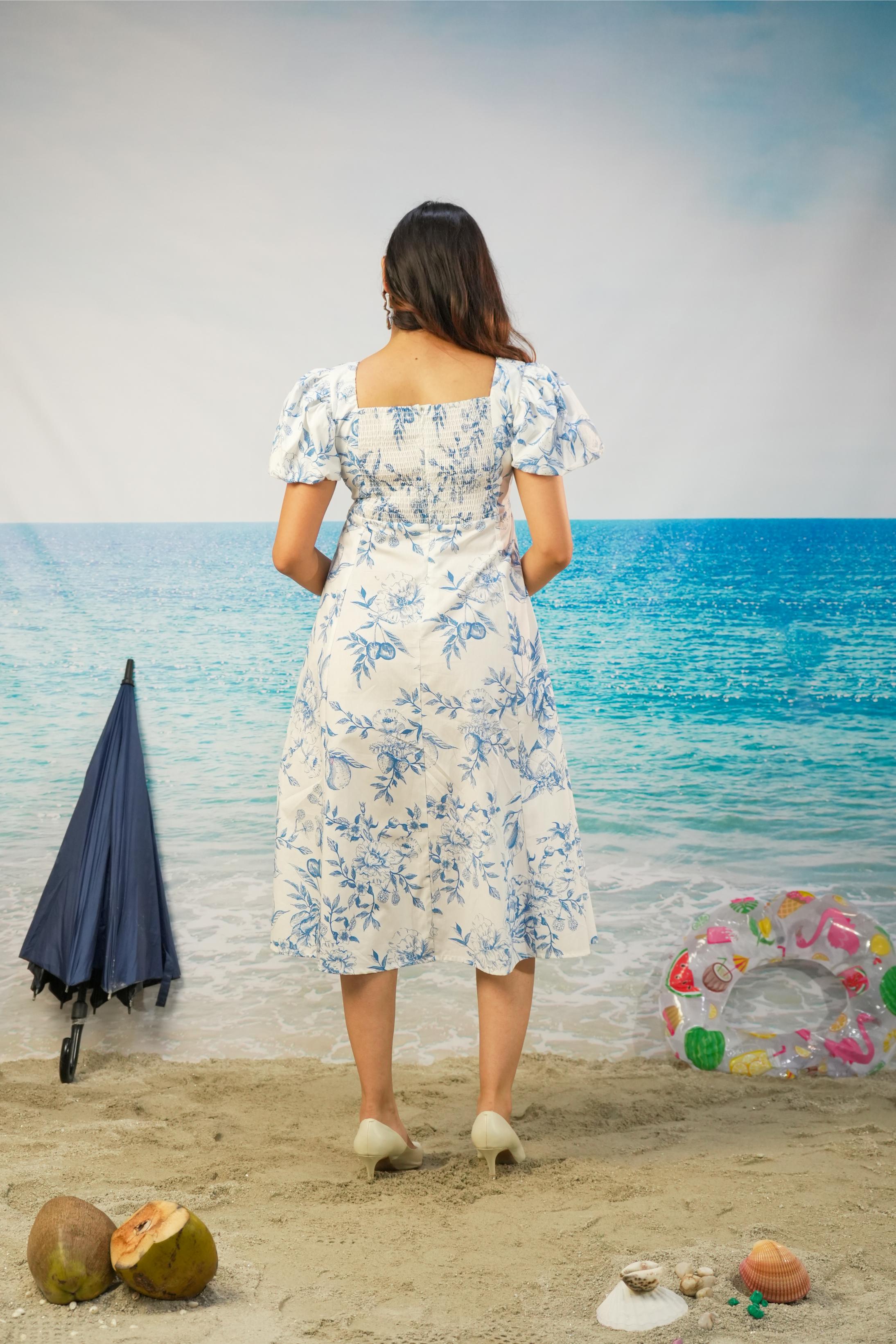 A woman stands with her back to the camera on a beach-themed backdrop, wearing a white midi dress with a blue floral pattern and puff sleeves. A closed navy umbrella, coconuts, and a colorful inflatable ring float on the sand beside her.