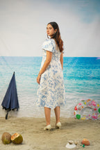 A woman stands on a beach backdrop wearing a white knee-length dress with a blue floral print. The scene includes sand, ocean water, a closed navy umbrella, a colorful inflatable ring buoy, and coconuts.
