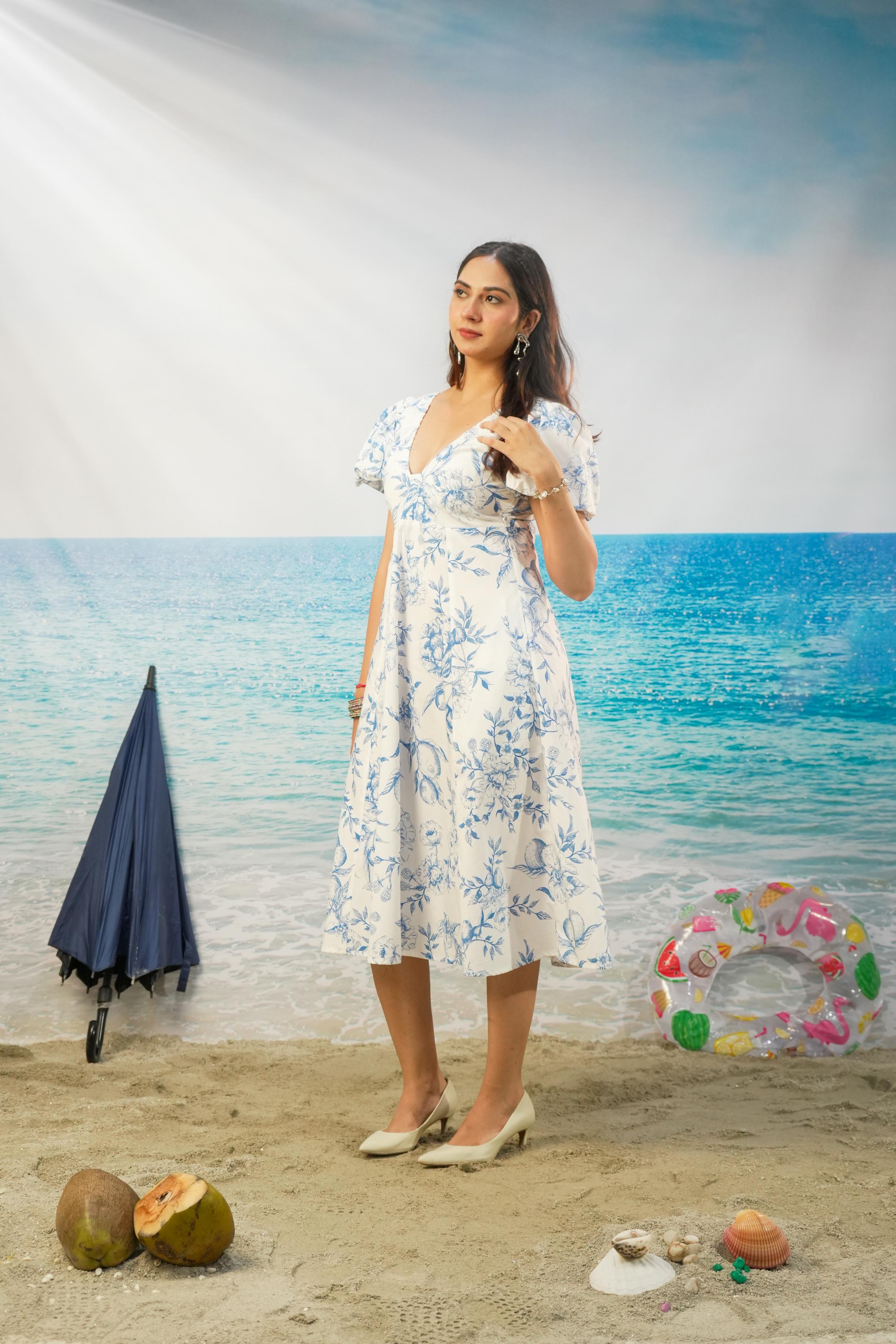 A woman stands on a beach backdrop wearing a white midi dress with a blue floral pattern. The scene includes sand, ocean, a closed navy umbrella, a colorful inflatable ring, and coconuts.