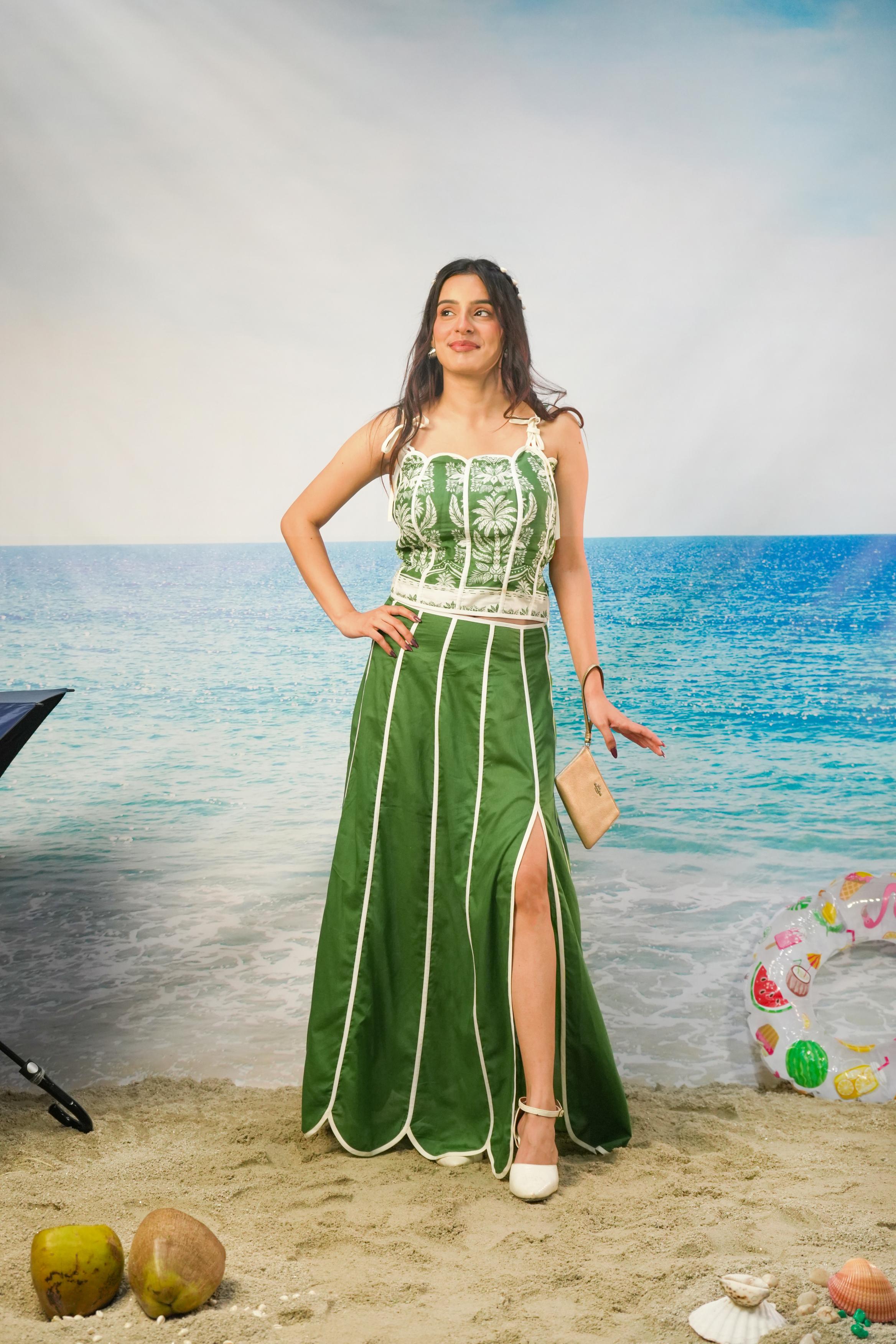 A woman modeling a gKavya Cotton Cambric Scalloped Co-ord Set with vertical white piping standing on a beach backdrop with ocean and sand visible behind her.