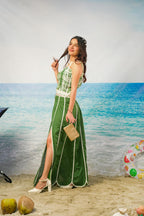 A woman stands on a beach backdrop wearing aKavya Cotton Cambric Scalloped Co-ord Set with white trim and a high slit, holding a small beige handbag. The background shows blue ocean water and pale sand, with a swim ring and coconuts visible near the bottom.