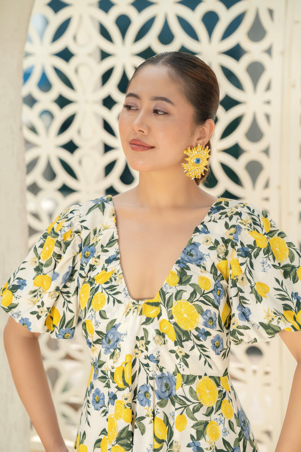 Woman wearing a Citrus Bloom Puff-Sleeve Dress in Cotton with lemon and blue flower pattern Zoom in Imgae against a decorative wall.