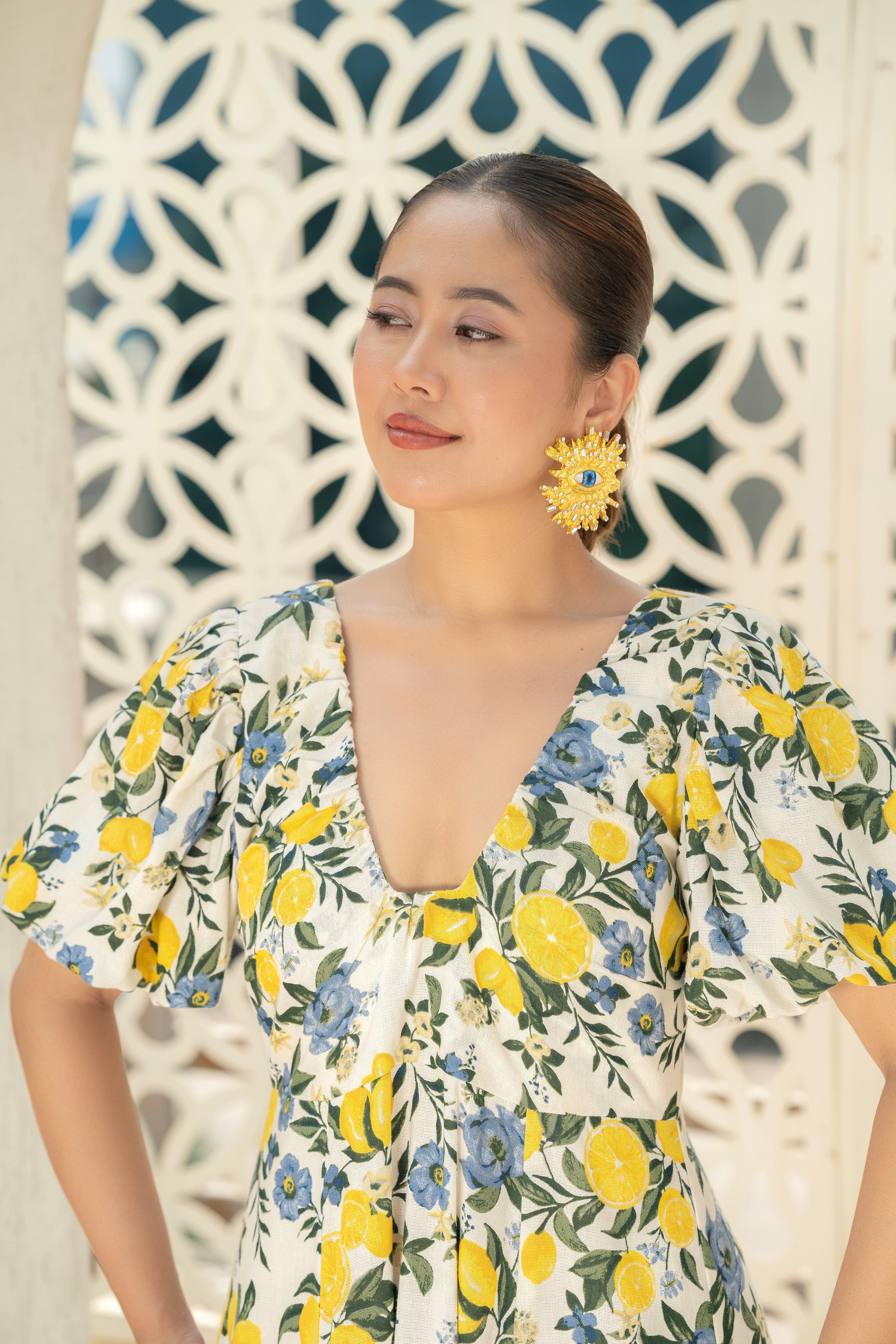 Woman wearing a Citrus Bloom Puff-Sleeve Dress in Cotton with lemon and blue flower pattern Zoom in Imgae against a decorative wall.