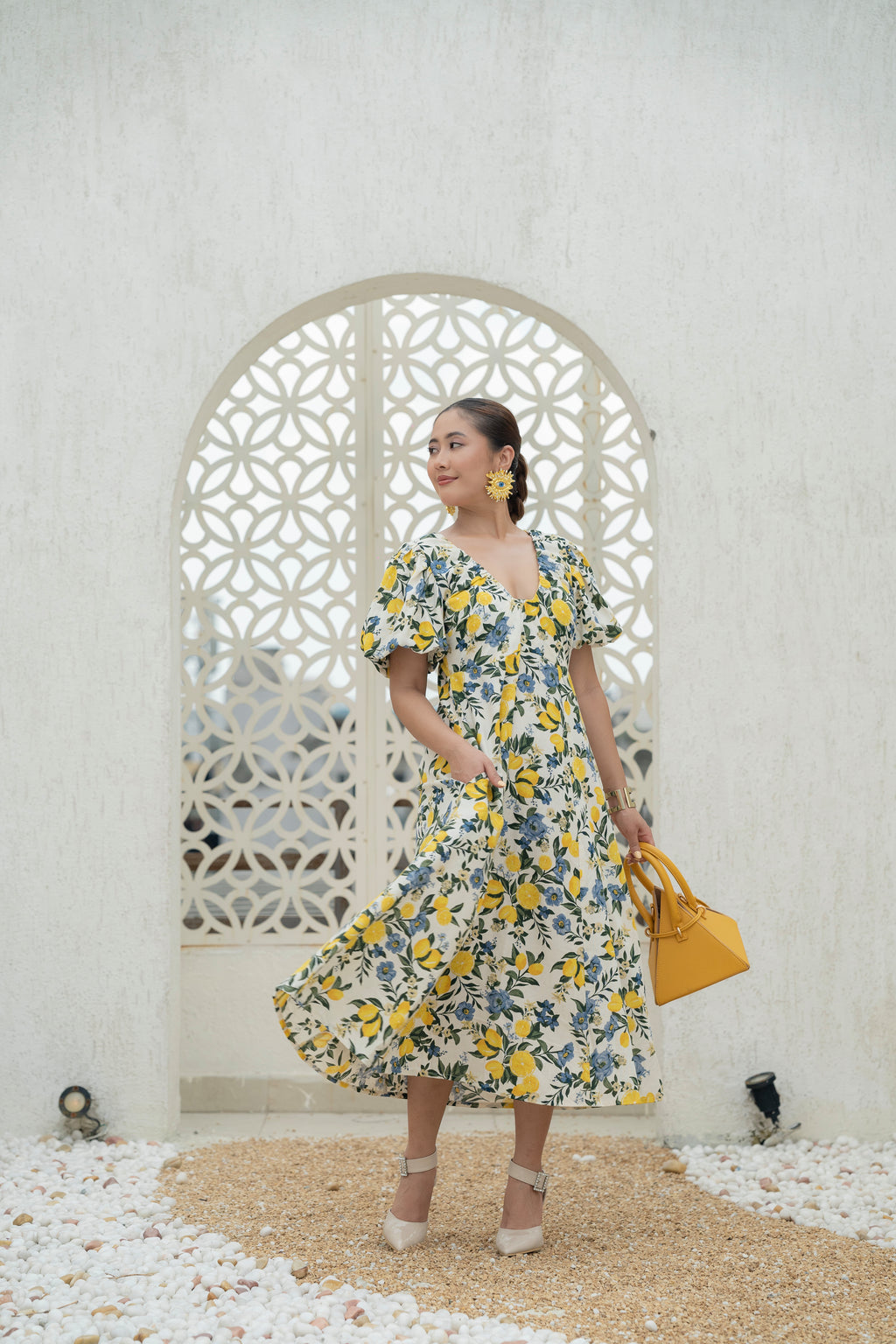 Woman in a Khamna's Citrus Bloom Puff Sleeve Dress in Cotton Flax Lemon and Floral Print holding a yellow bag in front of a decorative arch. Made for you Holiday, Vacation or Girls Brunch. Perfect for sunny days.