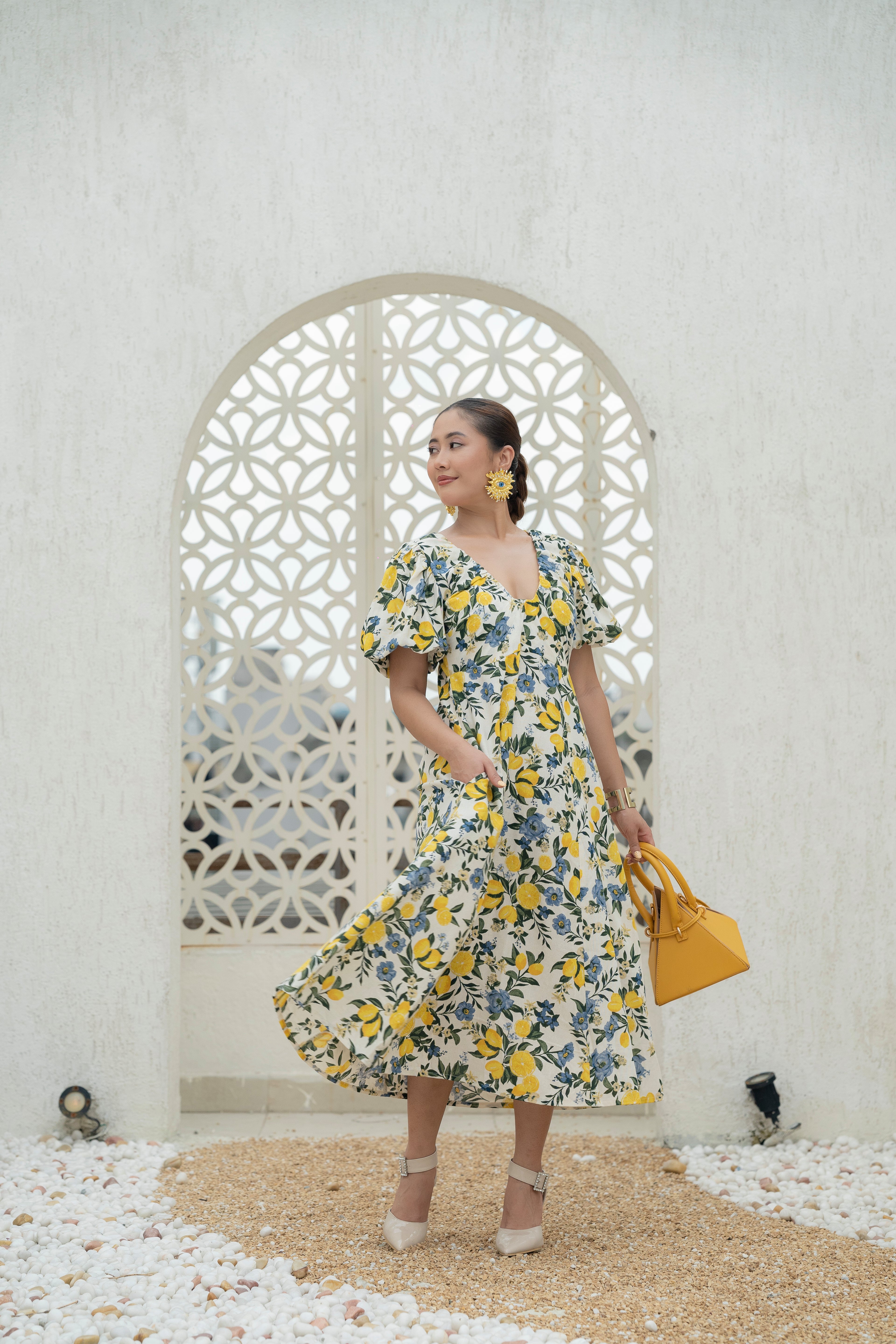 Woman in a Khamna's Citrus Bloom Puff Sleeve Dress in Cotton Flax Lemon and Floral Print holding a yellow bag in front of a decorative arch. Made for you Holiday, Vacation or Girls Brunch. Perfect for sunny days.