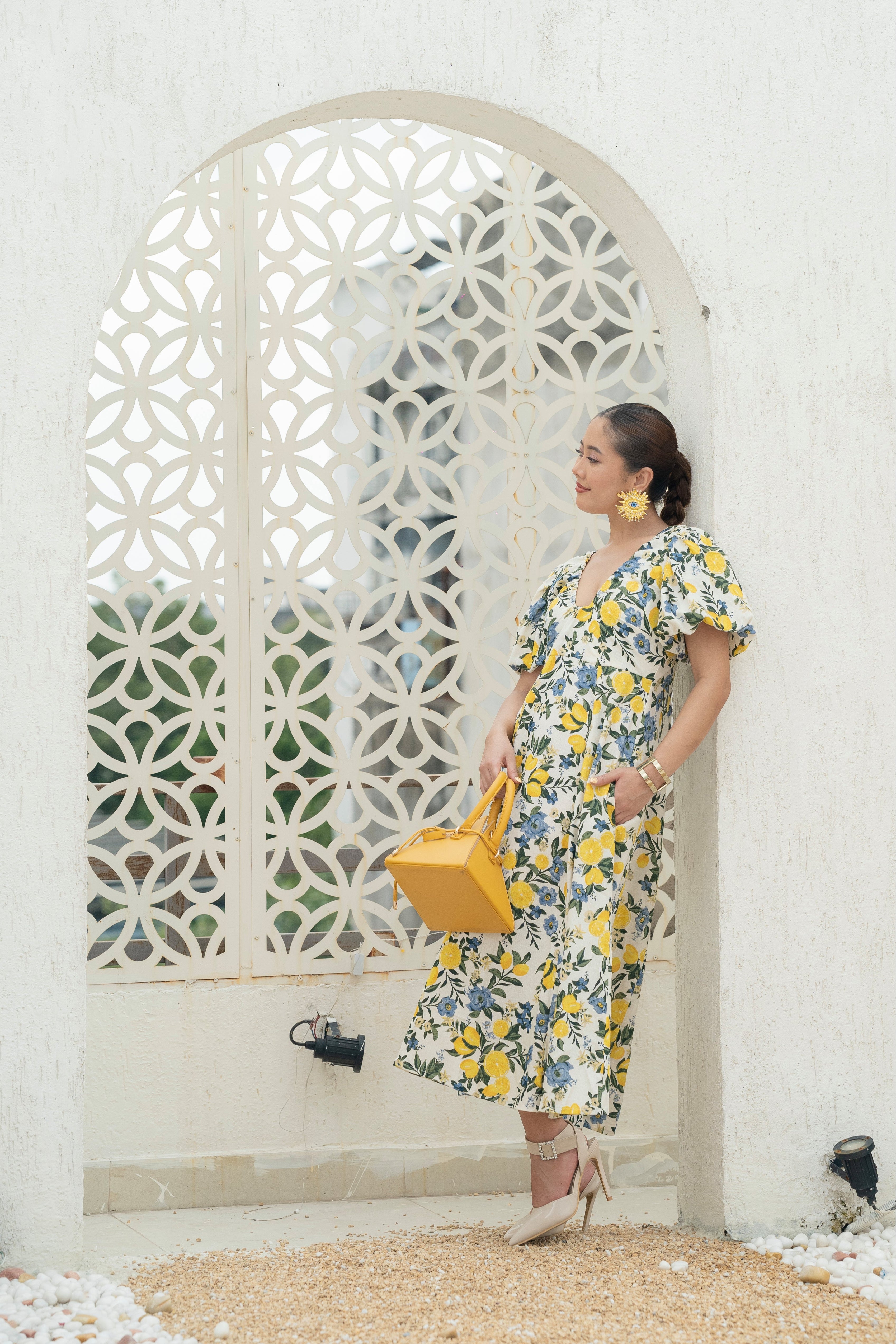 Woman in a Khamna Citrus Bloom Puff-Sleeve Dress in Cotton Flax, in lemon and Floral print Side look standing in front of a decorative white archway.
