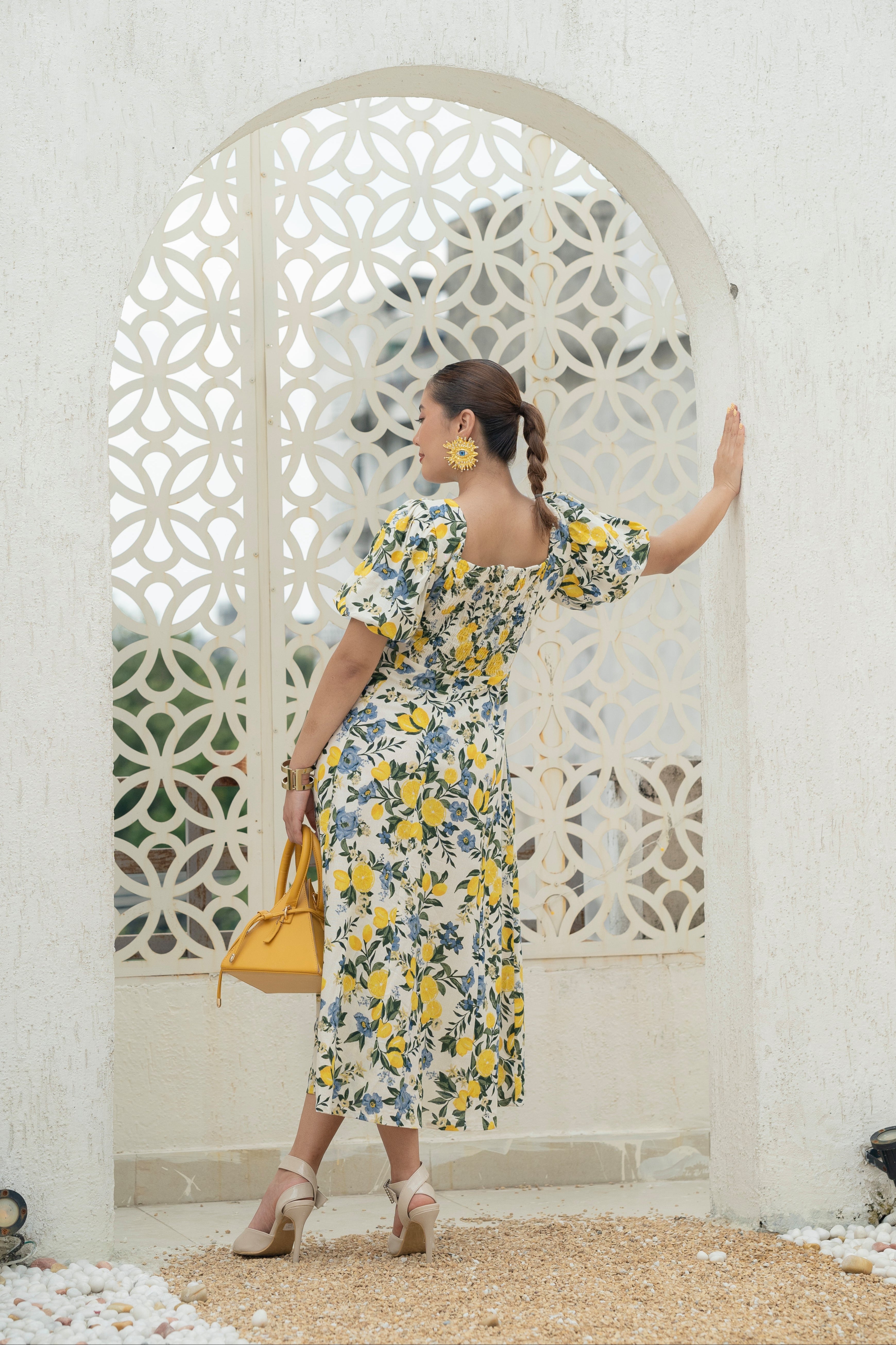 Woman in a Khamna's Citrus Bloom Puff-Sleeve Dress in Cotton Flax in Lemon and Floral print with Smocking at back in Lemon and Floral Print standing in front of a decorative white archway.