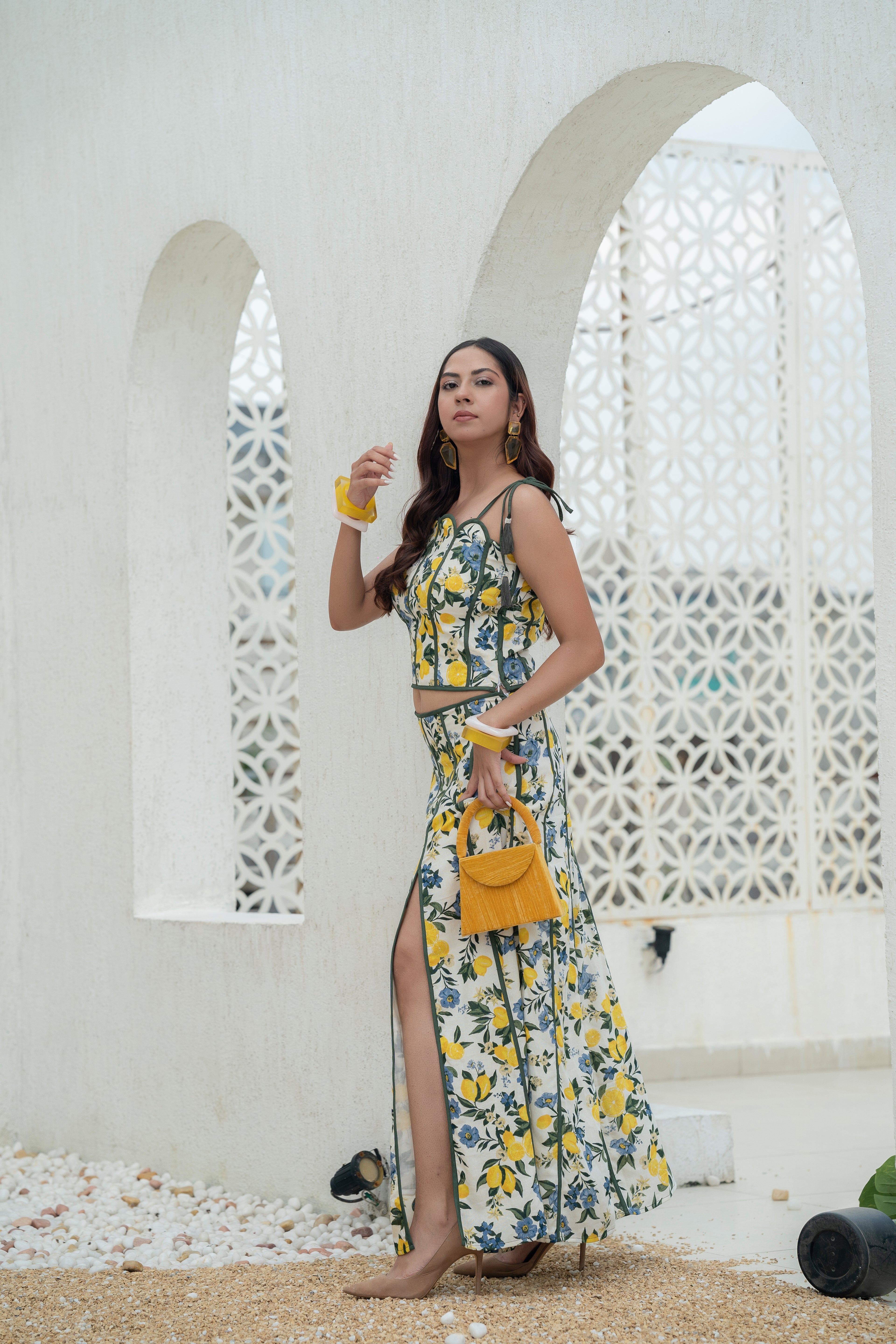 Woman in a floral Khamna Amalfi Bloom Skirt – Mid Waist Slit Maxi holding a yellow handbag against a white lattice background