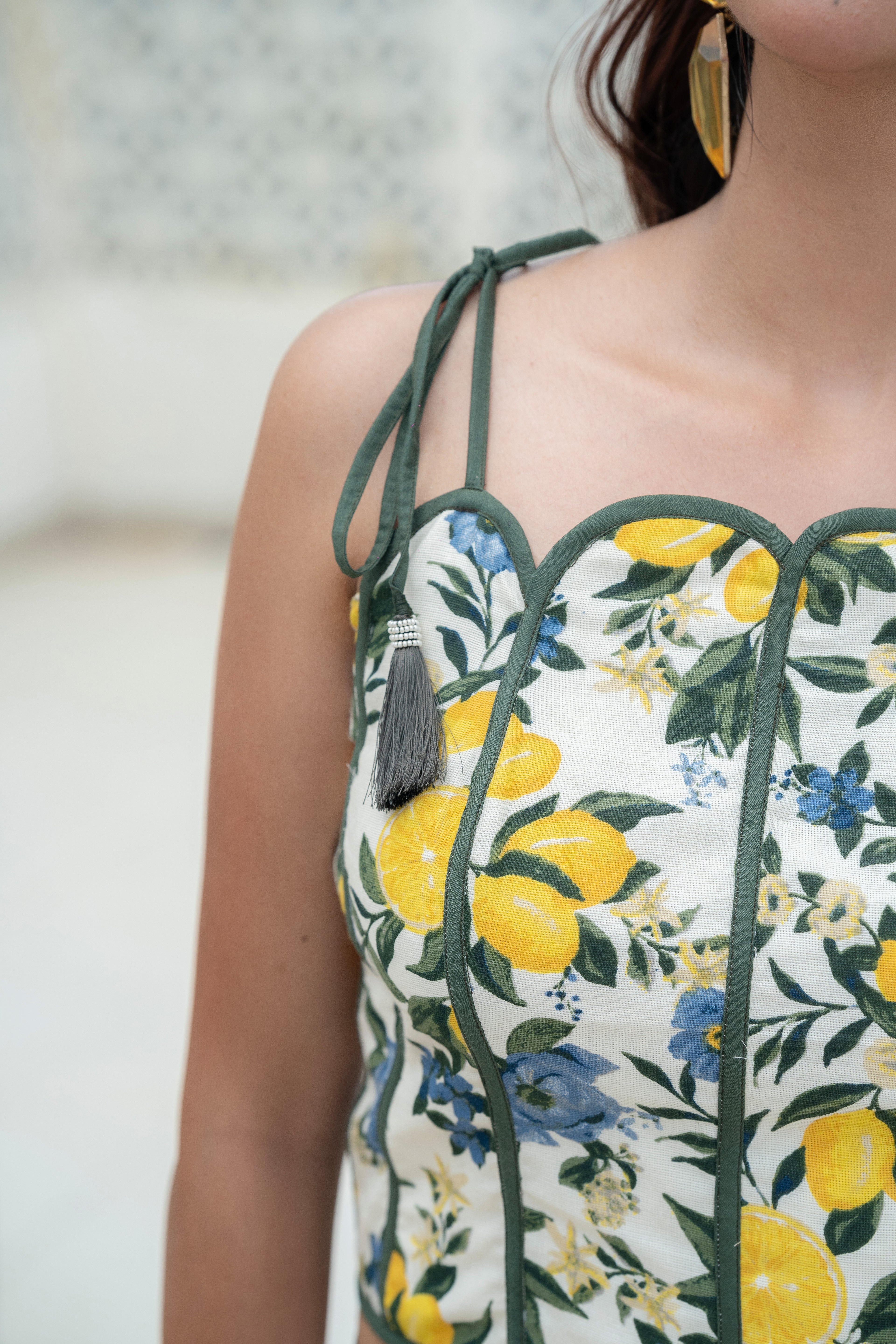 Close-up of a blue floral Khamna Amalfi Bloom Top – Scallop Detail Tassel Strap Crop with yellow lemons and green leaves on a blurred background
