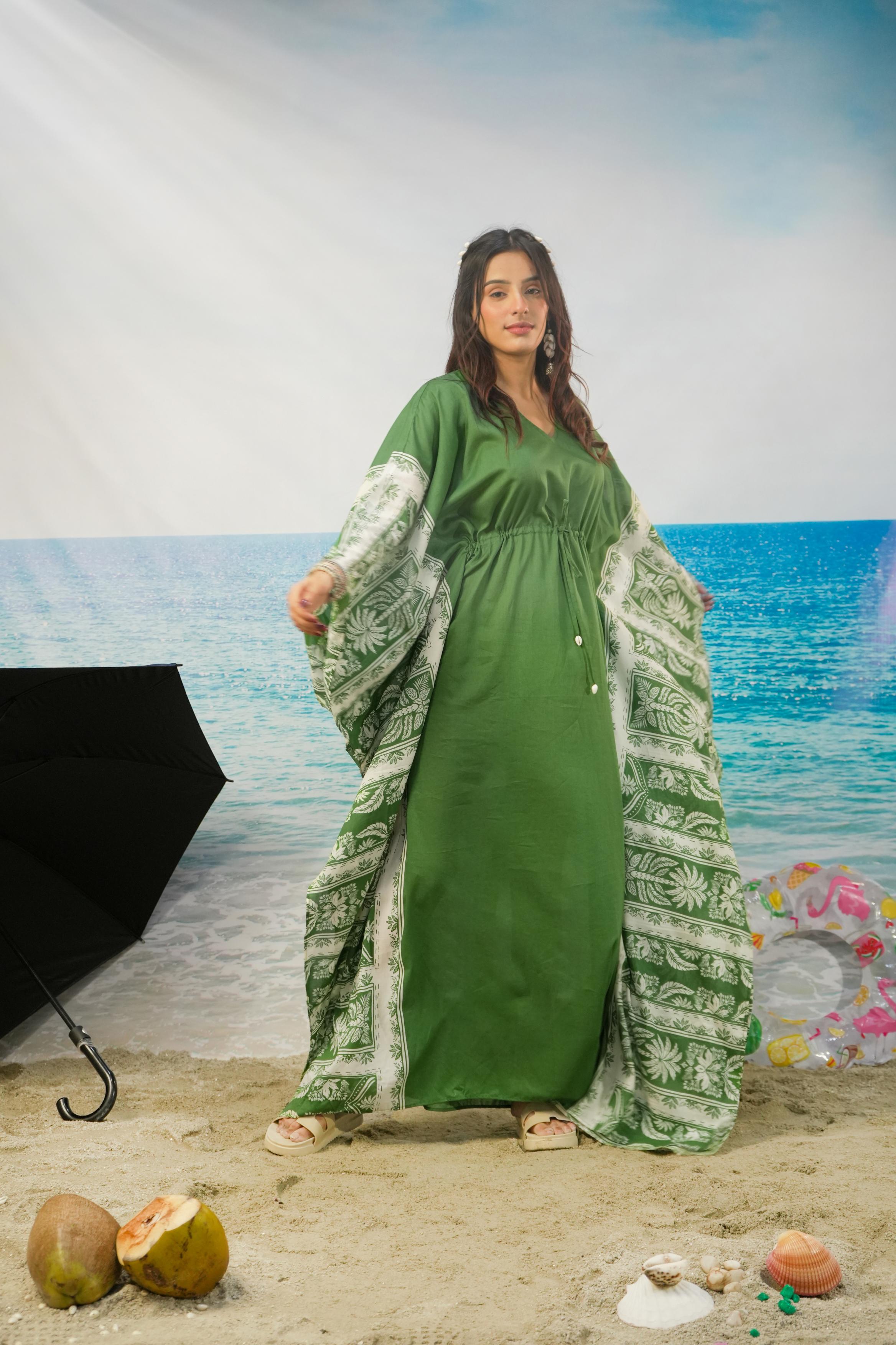 A woman modeling a green maxi dress with wide sleeves and a matching printed green-and-white shawl, posed on a sandy beach backdrop with ocean scenery behind her.