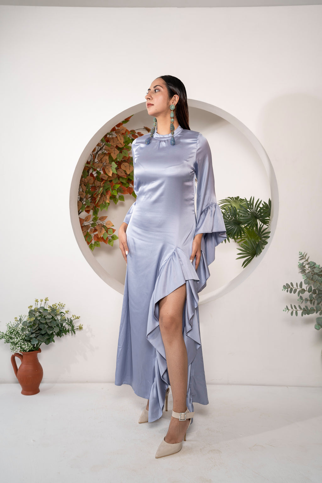 Woman in a Khamna Azure Muse - Stretch Satin Ruffle Gown standing in front of a circular plant display.