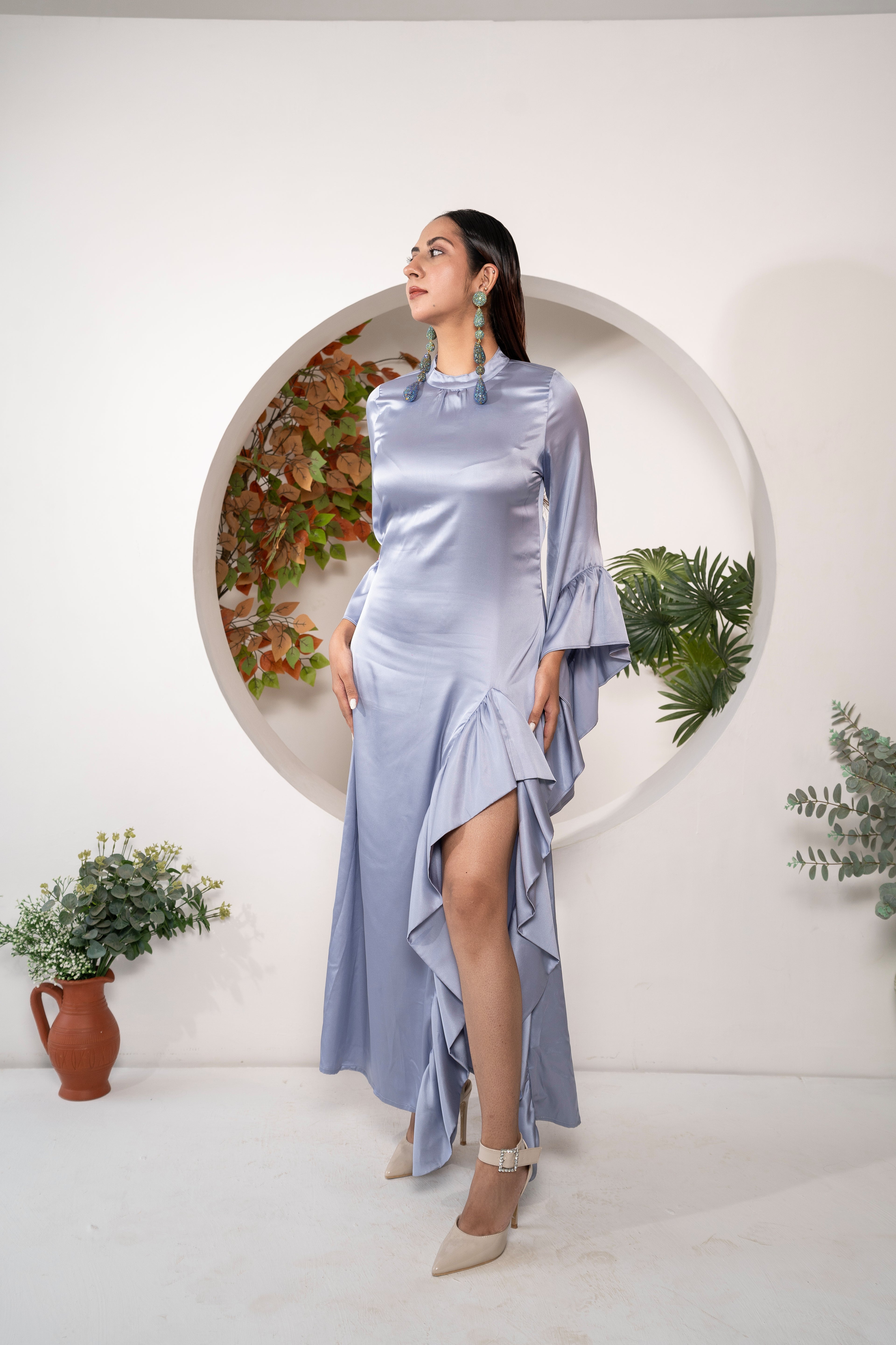 Woman in a Khamna Azure Muse - Stretch Satin Ruffle Gown standing in front of a circular plant display.