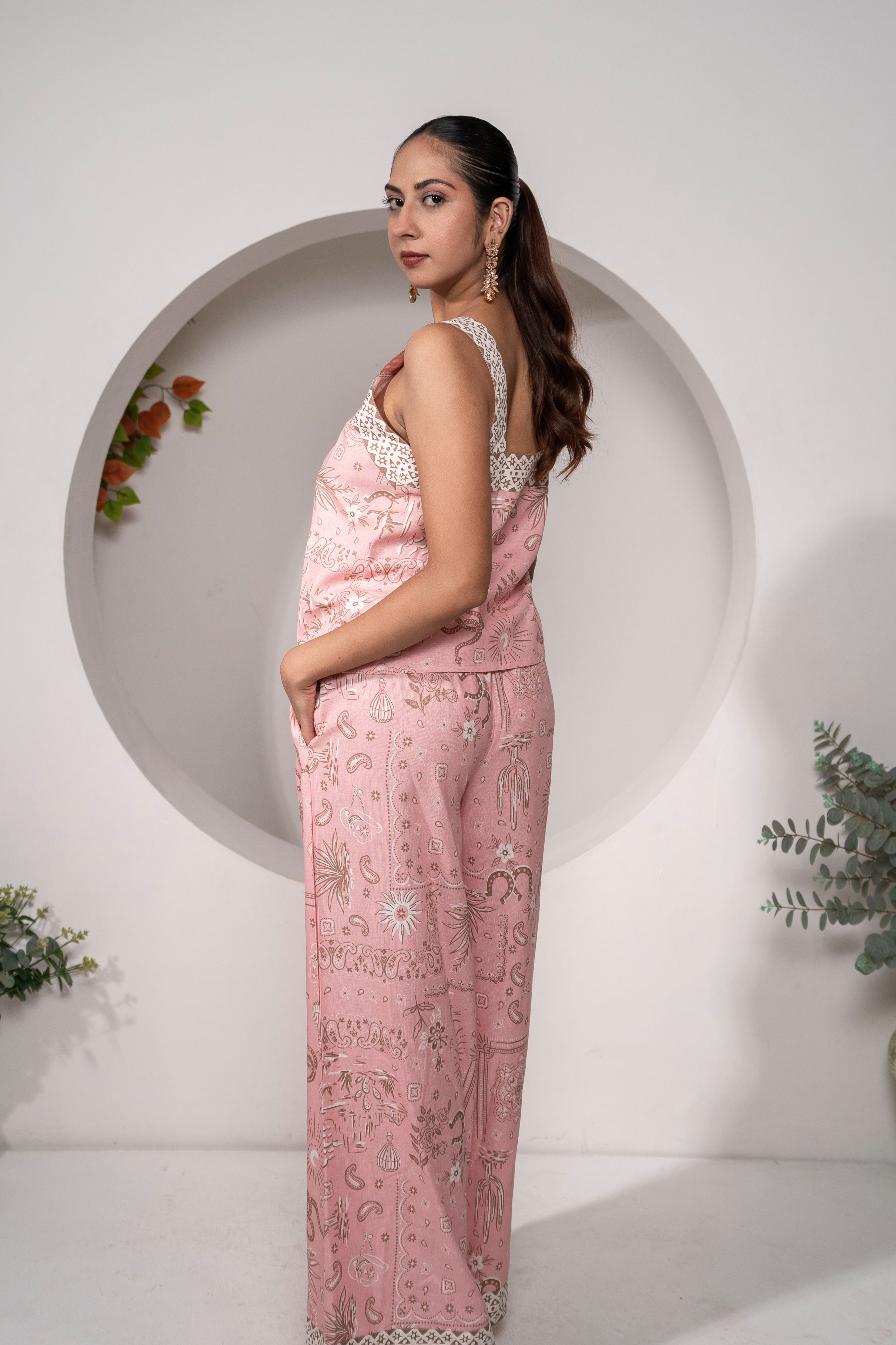 Woman wearing Khamna Desert Rose – Pink Printed Vacation Top Side View patterned outfit standing in front of a white wall with decorative elements.
