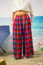 A model standing on a sandy beach wearing high-waisted, wide-leg plaid trousers with a vibrant red, pink, purple, and blue pattern, paired with a white cropped shirt.