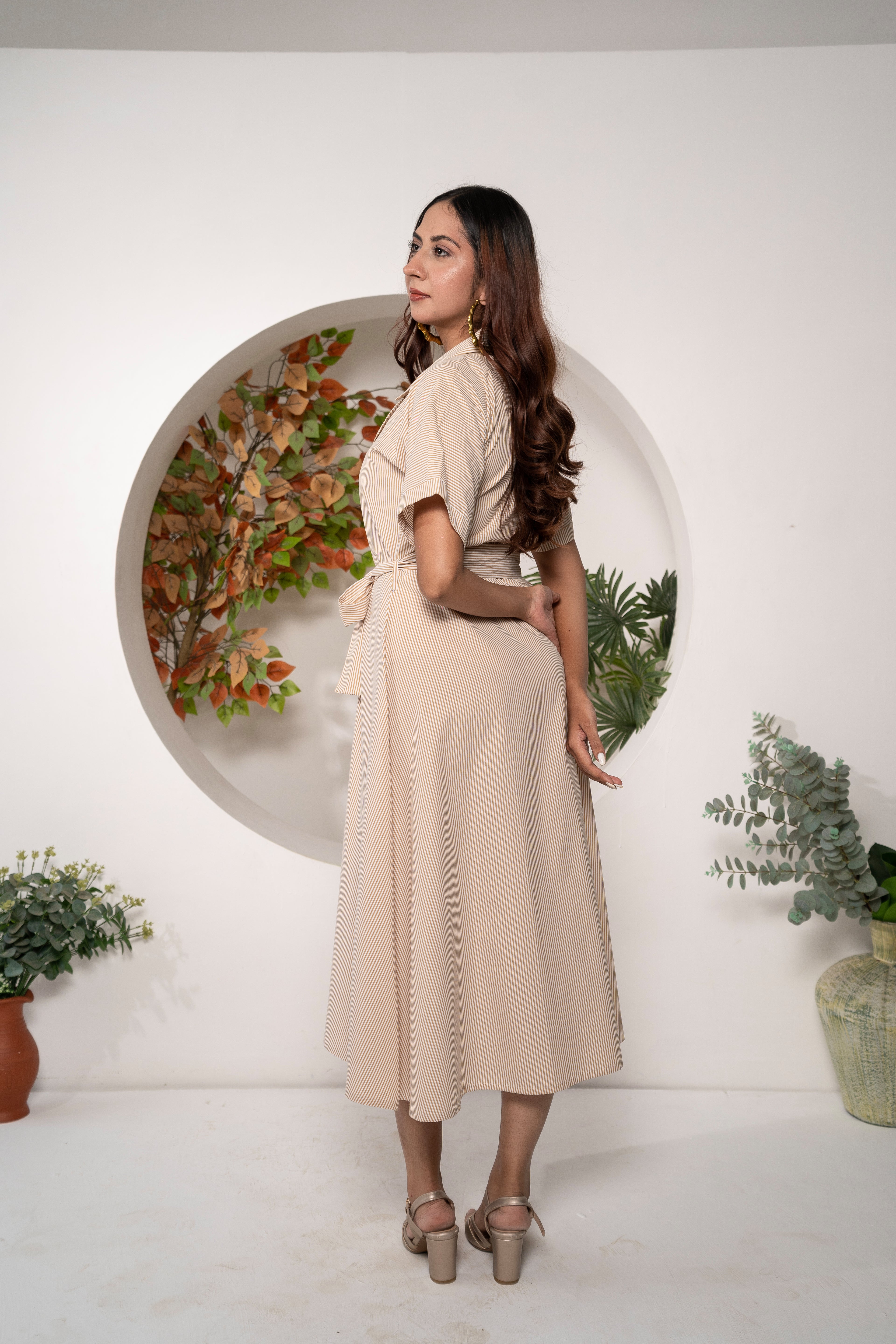 Woman in a Khamna Sandstone Stripe Shirt Dress – Cotton Y/D Collared Midi standing in front of a circular wall with floral design, surrounded by plants.