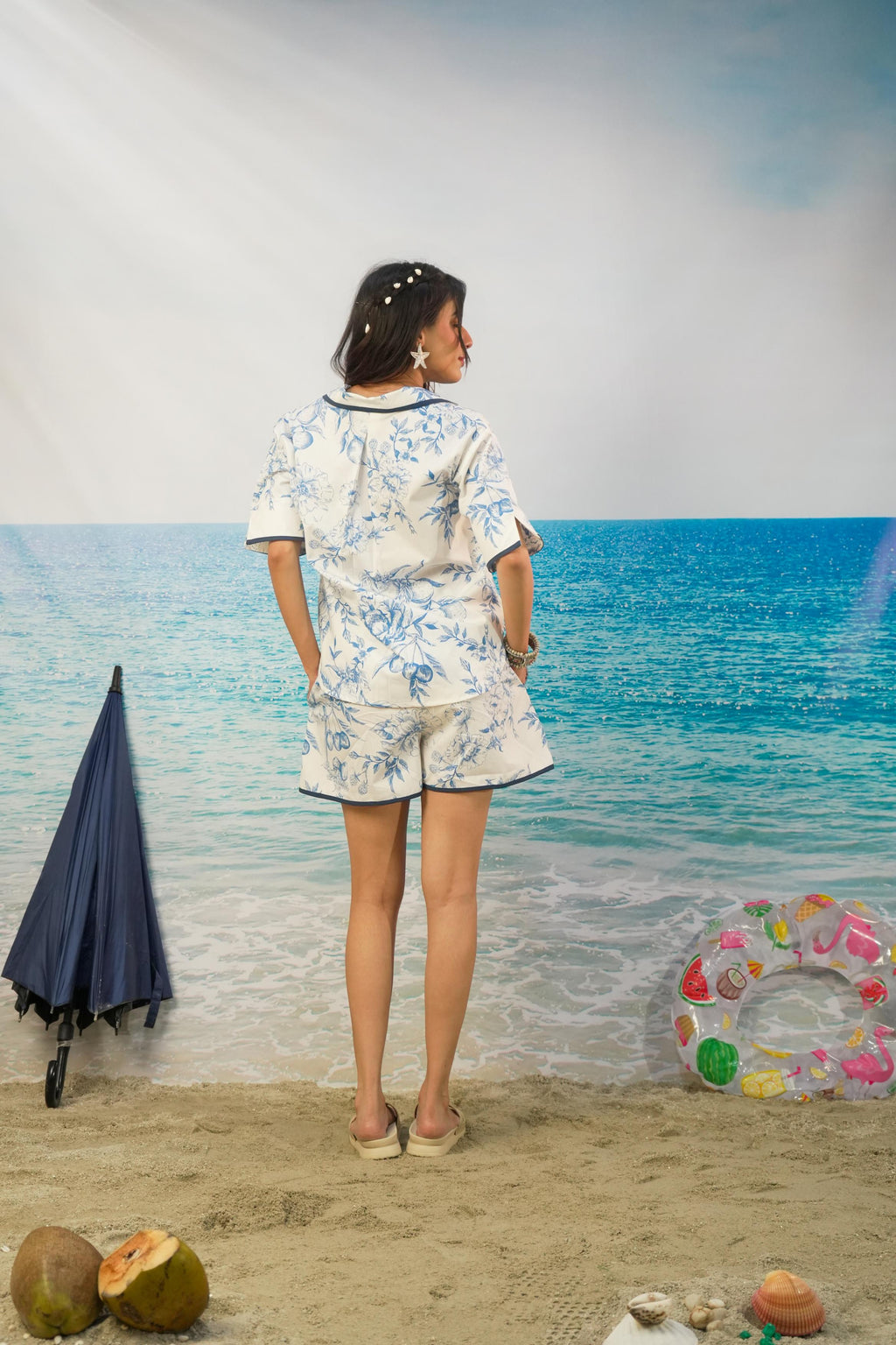 A woman stands on a beach backdrop wearing a matching short-sleeve top and shorts set in a white base with a blue tropical floral print. A closed navy umbrella, coconuts, and a pink swim ring float on the sand around her.