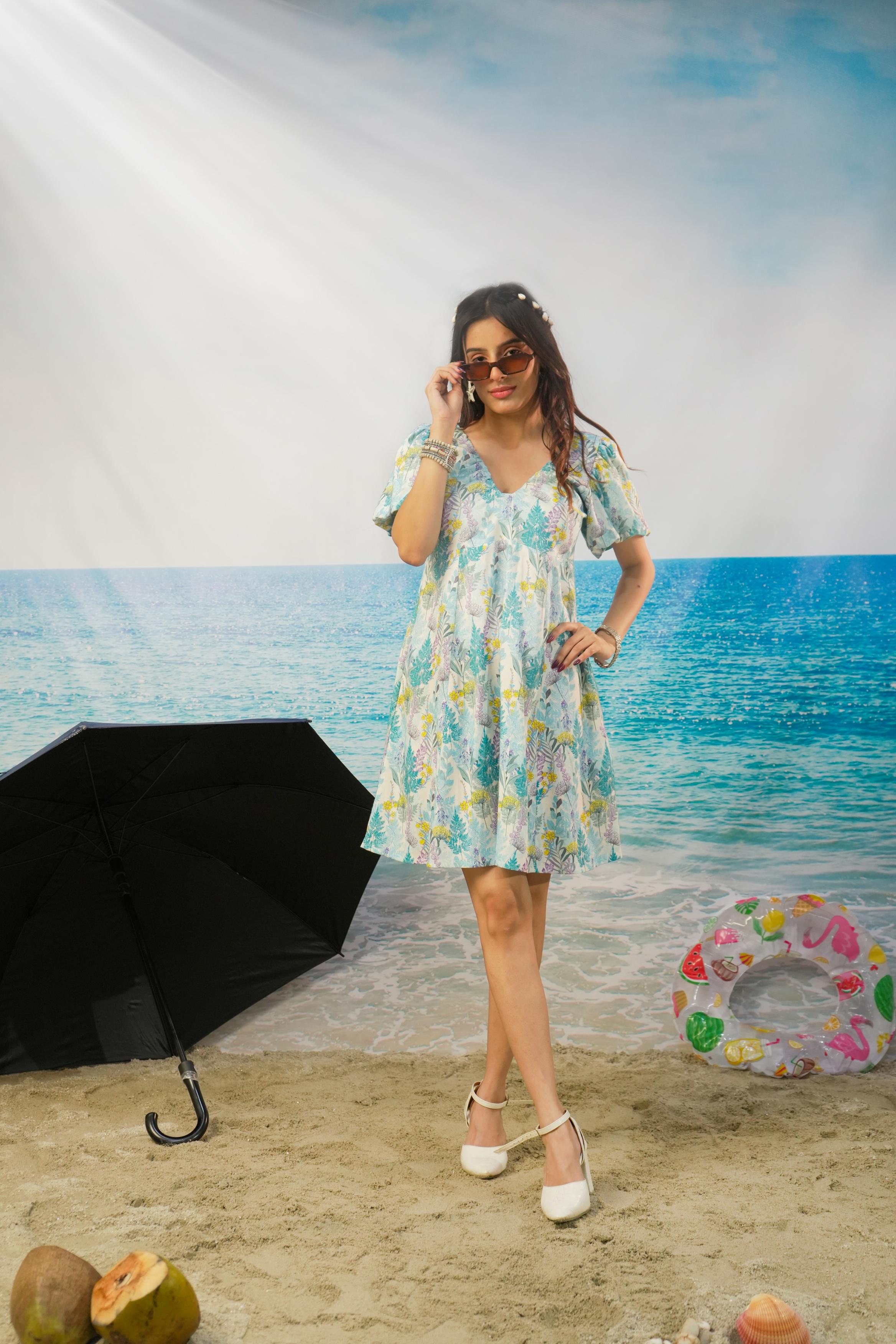 A woman modeling a Zohra Botanical Cotton Linen Puff Sleeve Dress | Short Resort Dress with a light blue, green, and yellow tropical floral print, standing on a sandy beach backdrop with a blue ocean and sky background. Props include a black umbrella, a pink and green inflatable ring, and coconuts on the sand.