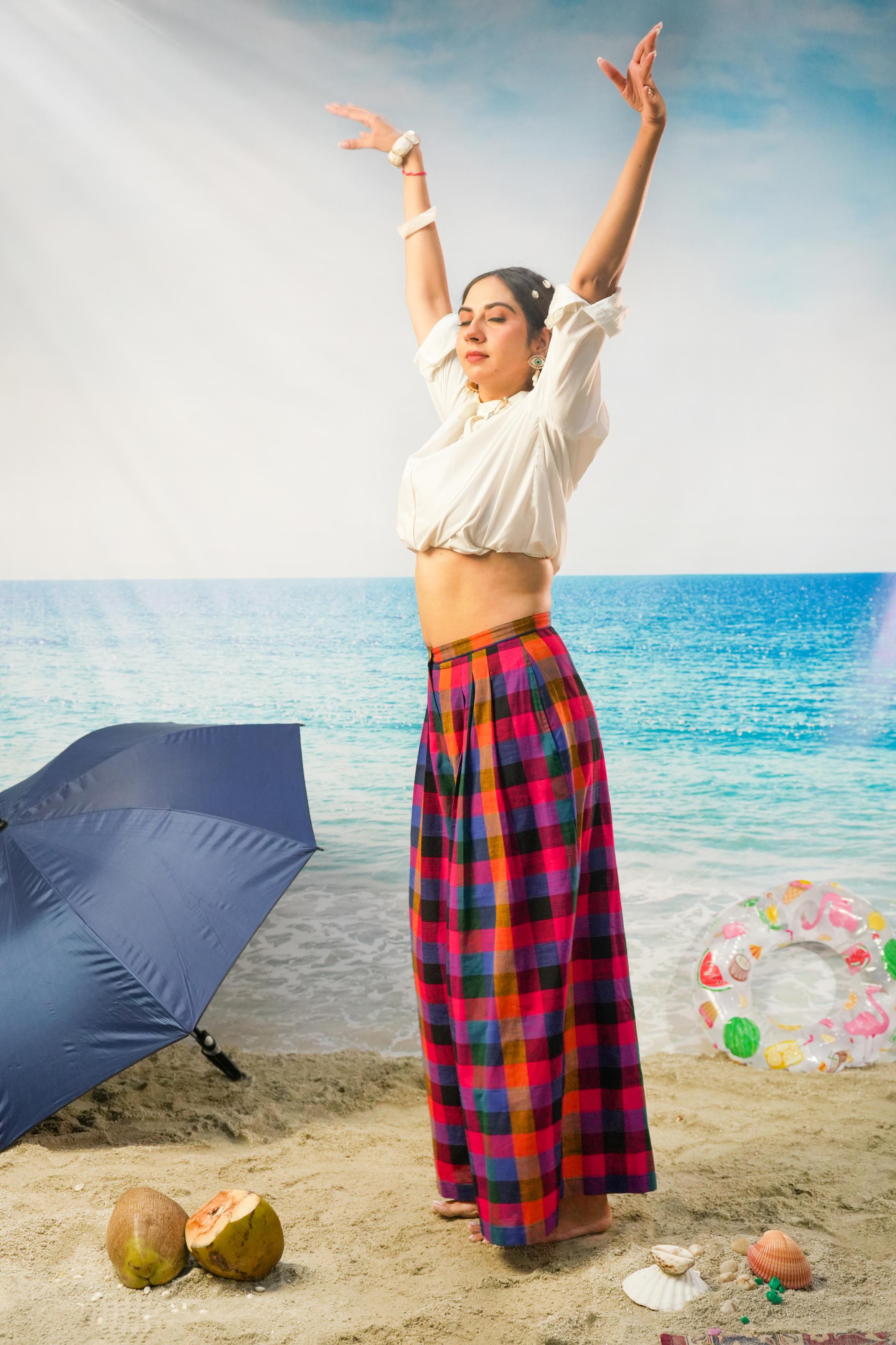 A woman stands on a beach with her arms raised, wearing a white cropped blouse and a colorful red, blue, and black plaid Pleated pants. A blue umbrella, coconuts, and a pool float are visible around her, with a beach and ocean backdrop.