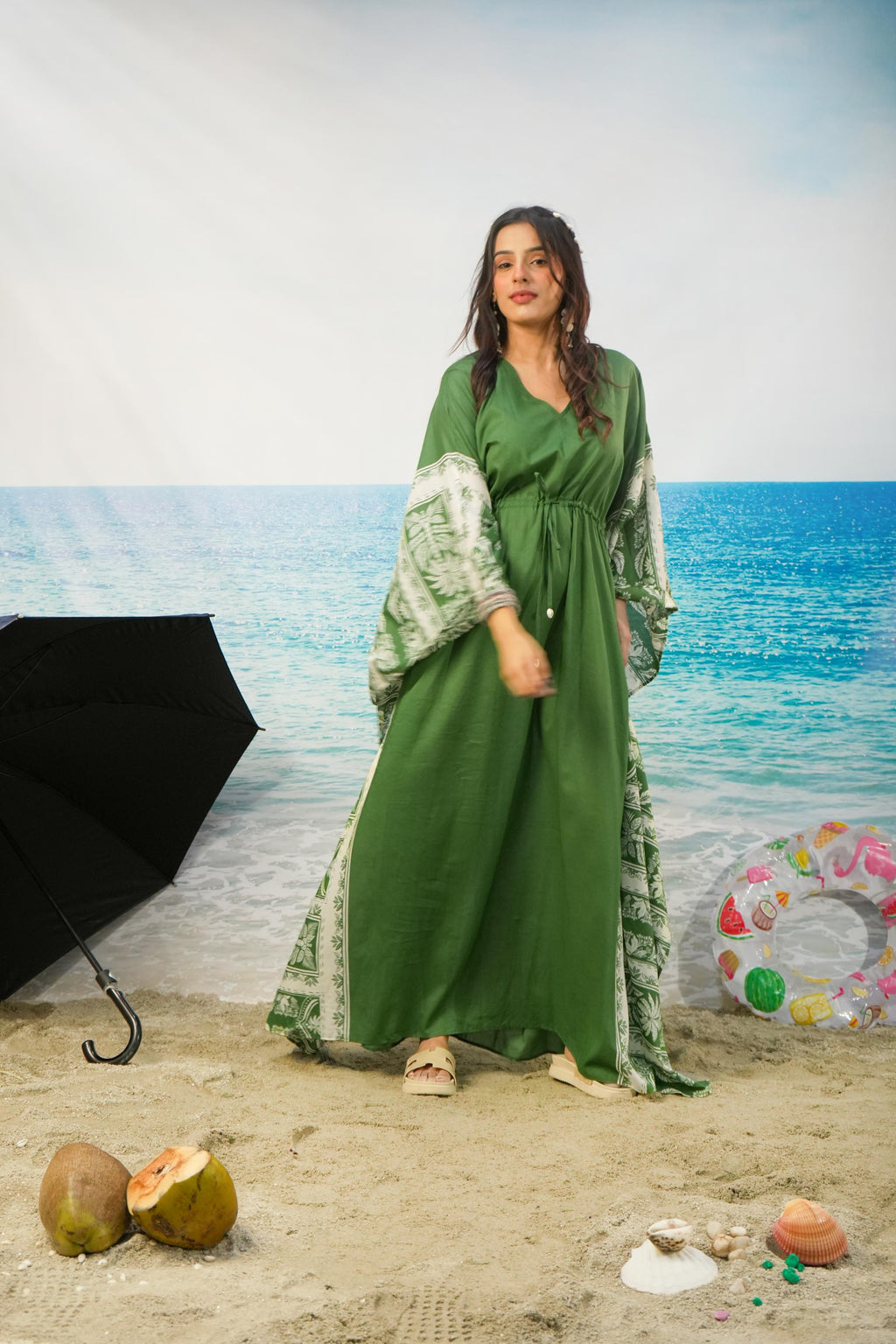 A woman modeling a long green kaftan dress with patterned sleeves and side panels, standing on a sandy beach backdrop with an ocean scene behind her. Decorative props include a black umbrella, coconuts, shells, and a colorful float.