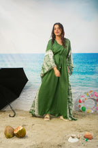 A woman modeling a long green kaftan dress with patterned sleeves and side panels, standing on a sandy beach backdrop with an ocean scene behind her. Decorative props include a black umbrella, coconuts, shells, and a colorful float.
