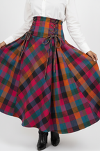 A person wearing a high-waisted, flared tartan plaid skirt in pink, orange, green, and black, paired with a white blouse and black boots, shown against a light gray studio background.