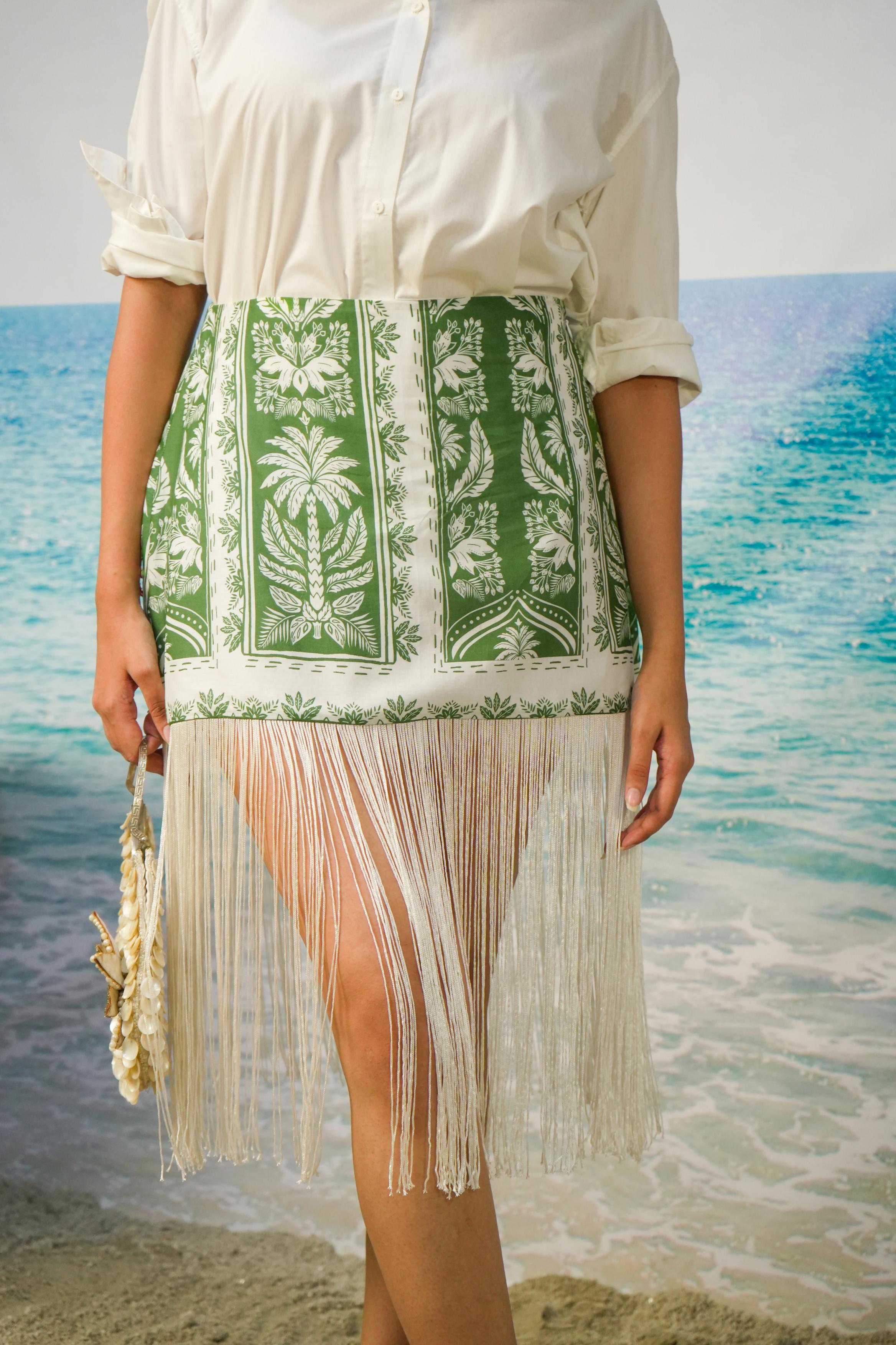 A woman wearing a white button-up shirt and aNaina Fringe Skirt | Green Botanical Resort Skirt with Fringe Hem, standing on a beach with turquoise water in the background.