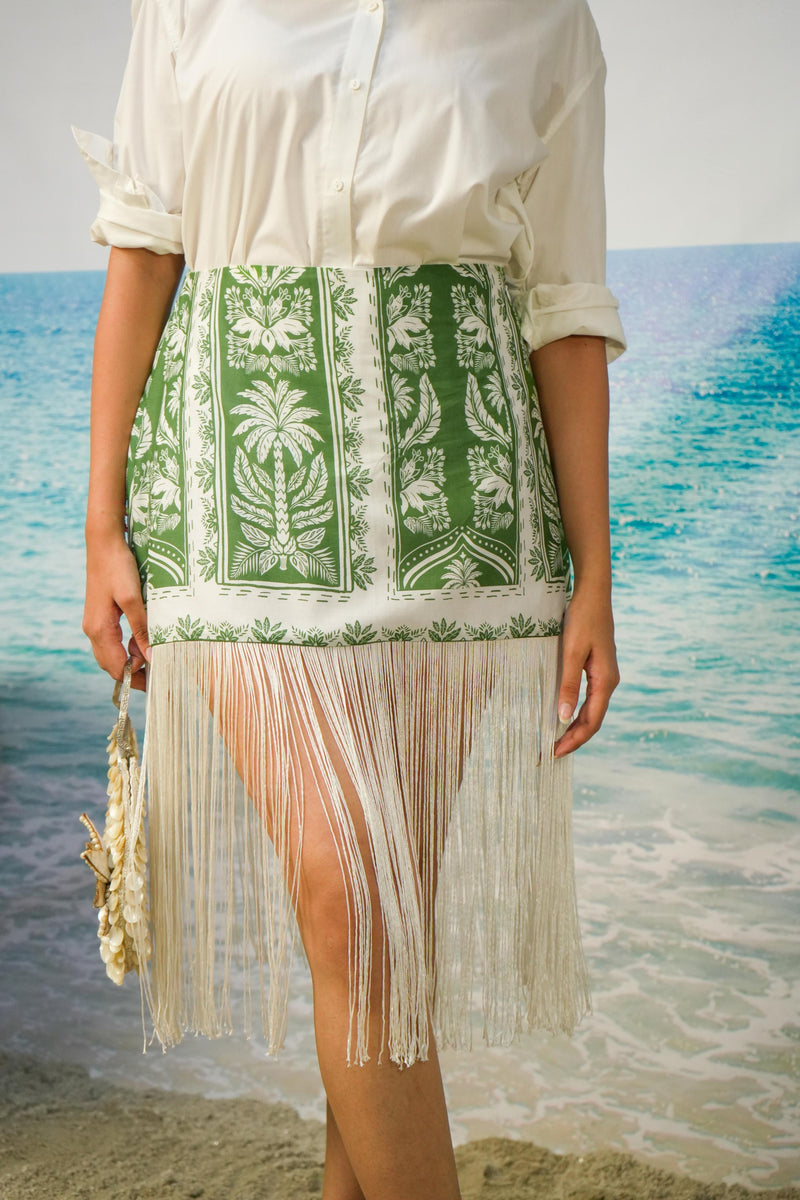 A woman wearing a white button-up shirt and aNaina Fringe Skirt | Green Botanical Resort Skirt with Fringe Hem, standing on a beach with turquoise water in the background.