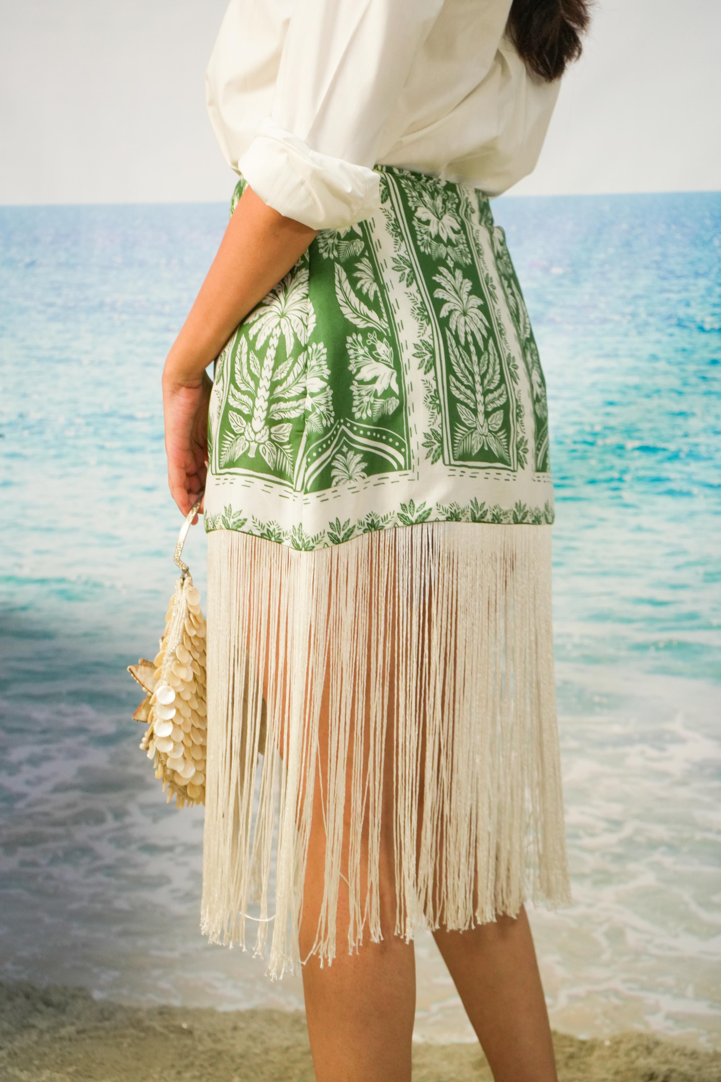 A woman standing on a beach wearing Naina Fringe Skirt | Green Botanical Resort Skirt with Fringe Hem, holding a woven straw bag.